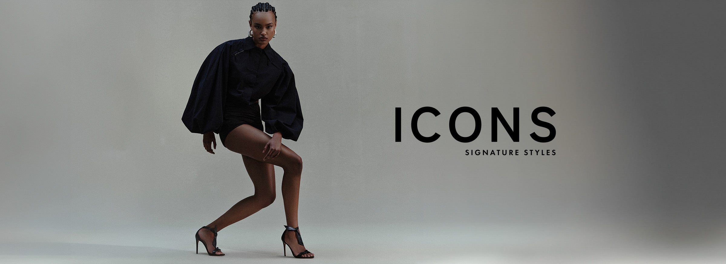 A fashion-forward woman dons chic black hot pants and a puffy sleeve black shirt, complemented by the classic Clarita 75 Sandal in Black. Text reads: Icons: Signature Styles.