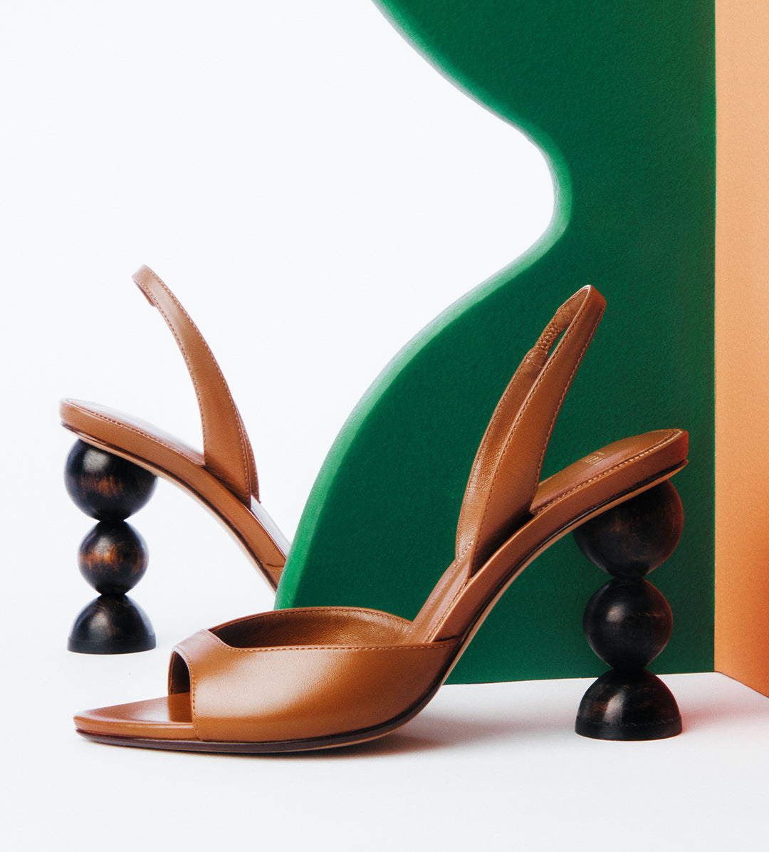 A pair of tan leather slingback heels featuring architectural heels composed of three stacked dark wooden spheres, set against green and orange geometric shapes.