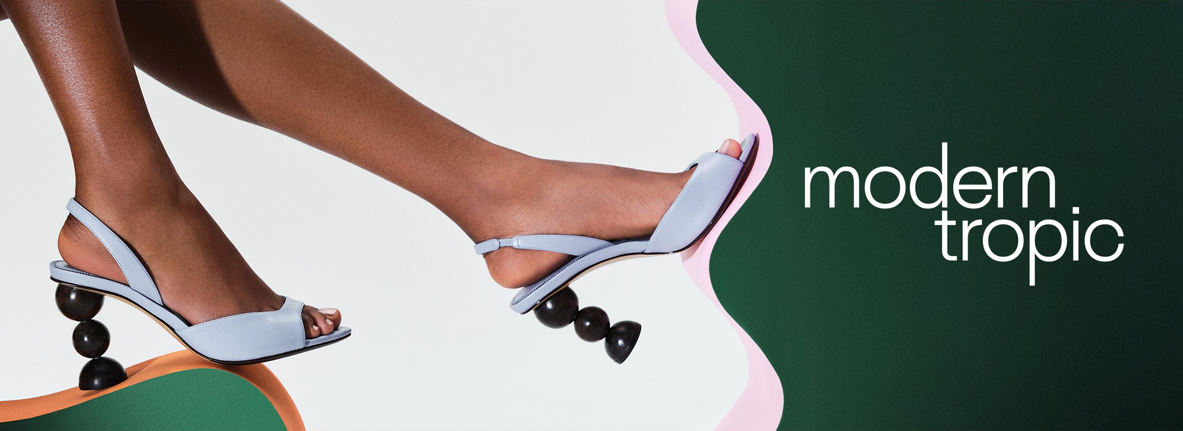  Close-up of a model's foot wearing a light blue leather slingback pump with a unique sculptural heel made of three stacked dark wooden spheres, against a green and pink abstract background with the text "MODERN TROPIC".