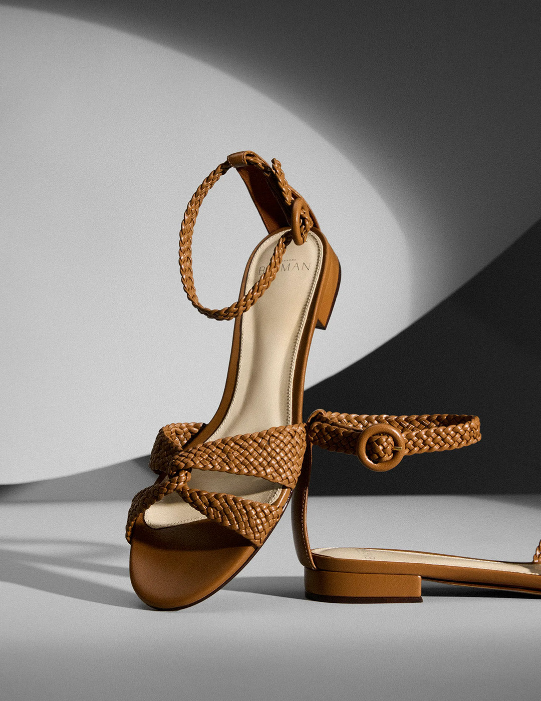 Brown heeled sandals with braided straps, one standing upright and the other leaning against it, on a smooth surface with dramatic lighting. Tap to shop