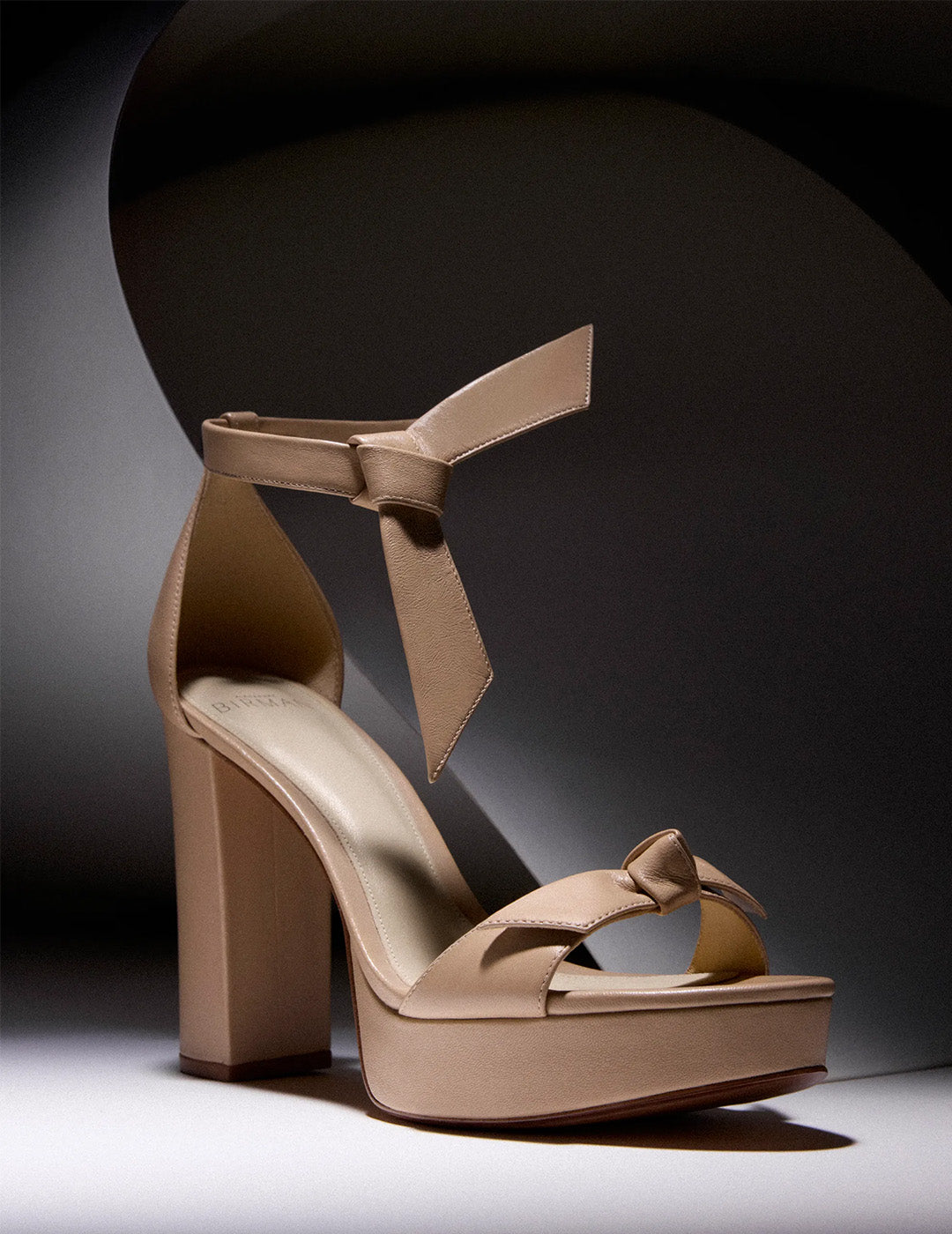 Elegant tan high-heeled sandal with ankle strap and knot accents on toe and ankle. Set against a shadowy, minimalist background, exuding sophistication.