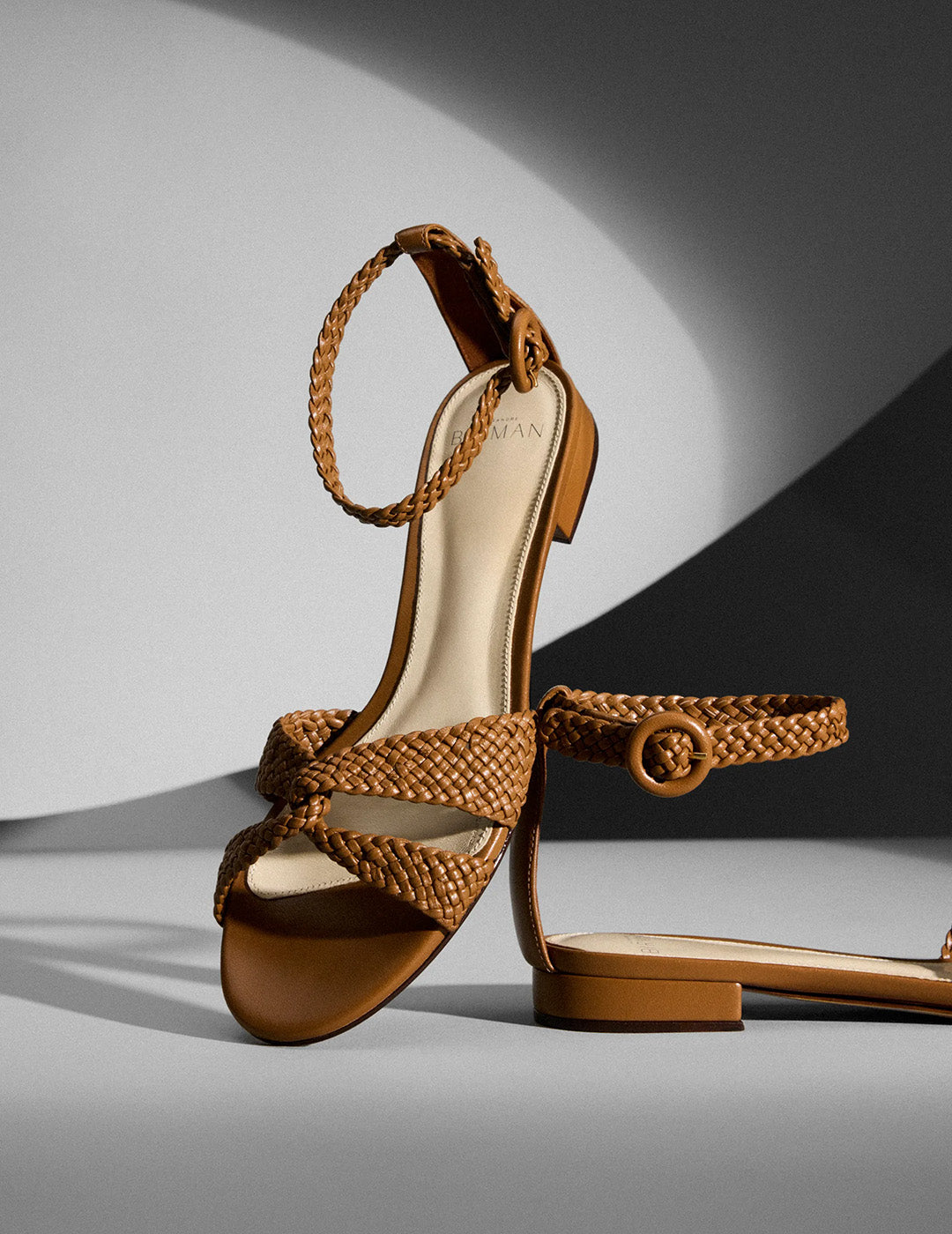 Stylish brown woven sandals with ankle straps, one upright and one laid flat, on a gray background. The lighting creates soft shadows and an elegant tone.