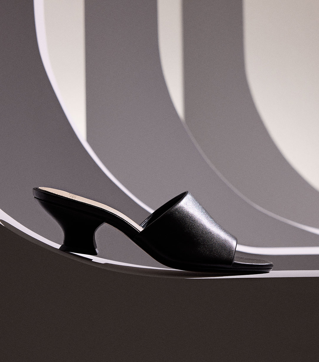 A black mule with a flared heel is positioned against a background of curved gray and white stripes. Tap to shop New Sandals.