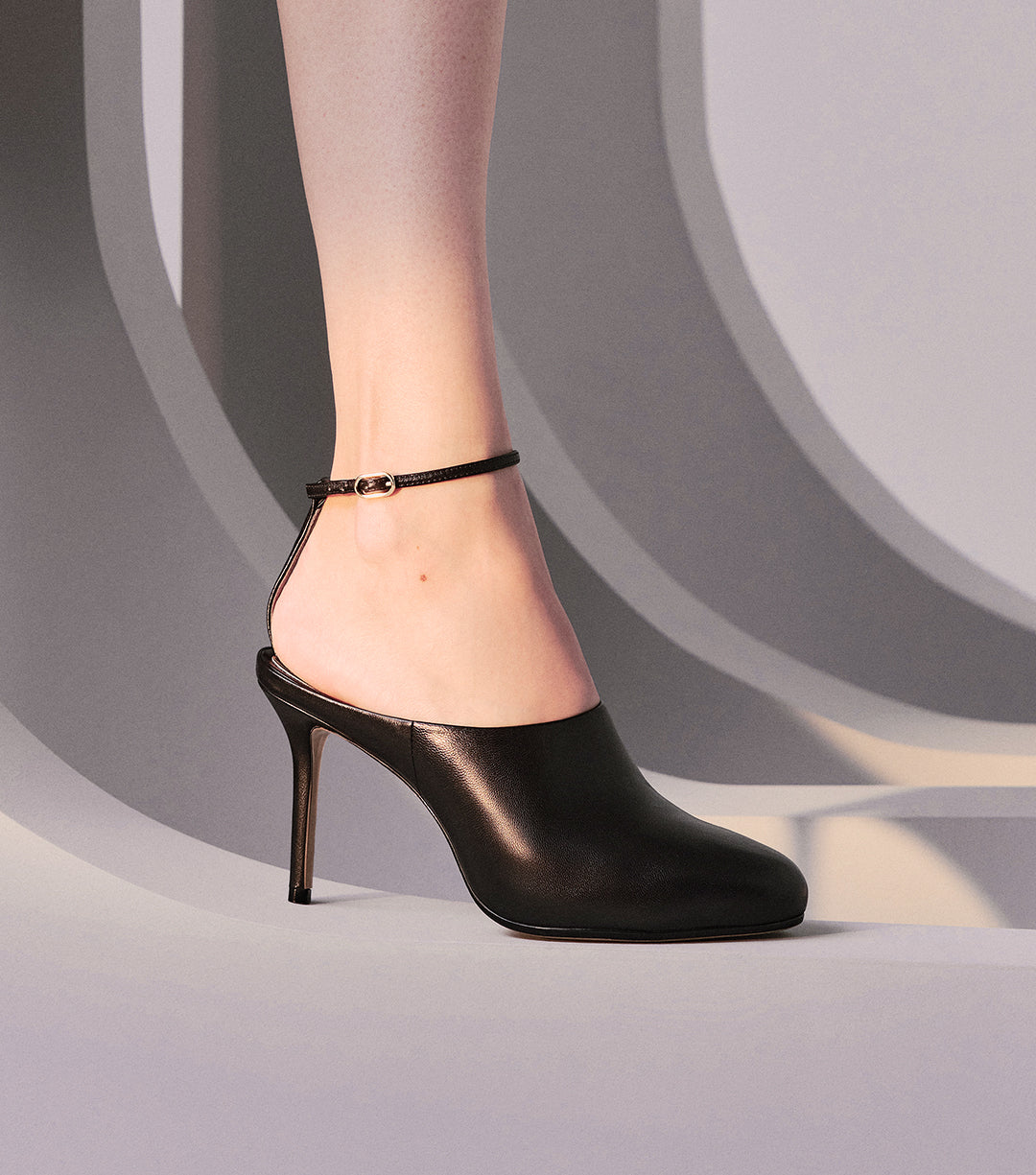 A person's foot is shown wearing a black pump-style mule with a stiletto heel and an ankle strap, against a background of gray and white stripes. Tap to shop New Pumps.