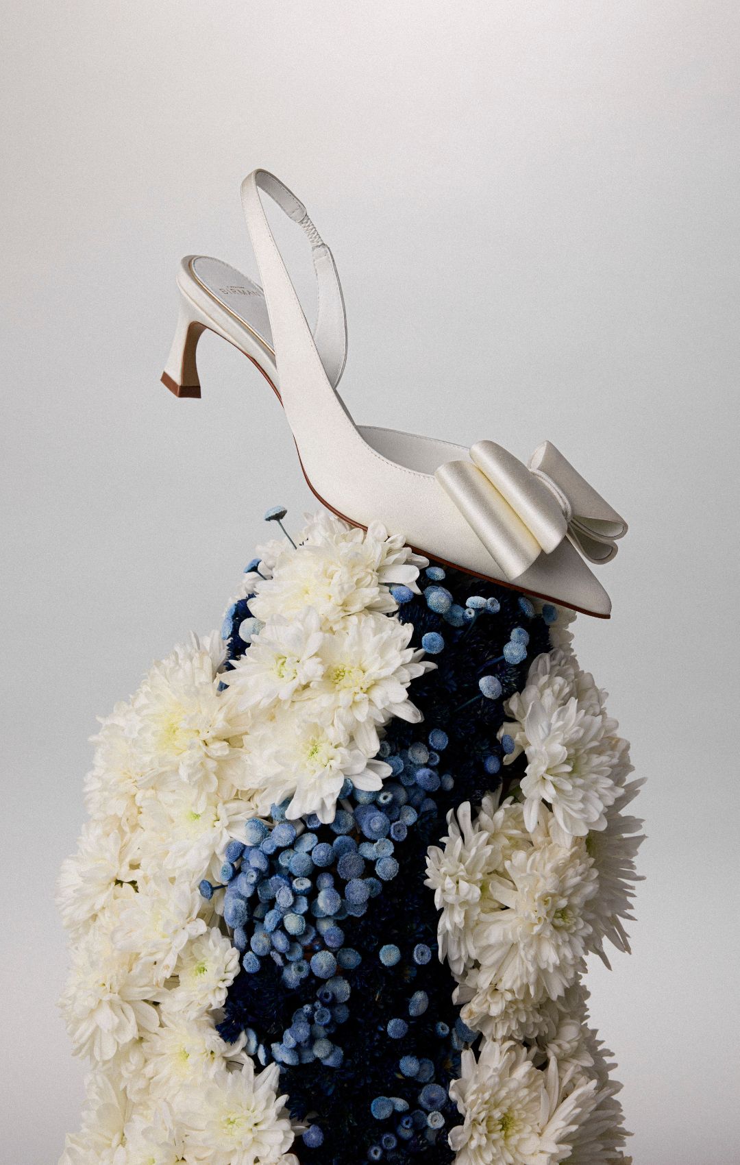  White high-heeled sandal rests atop a lush floral display of white chrysanthemums and blue berries, creating a whimsical, elegant fashion scene.