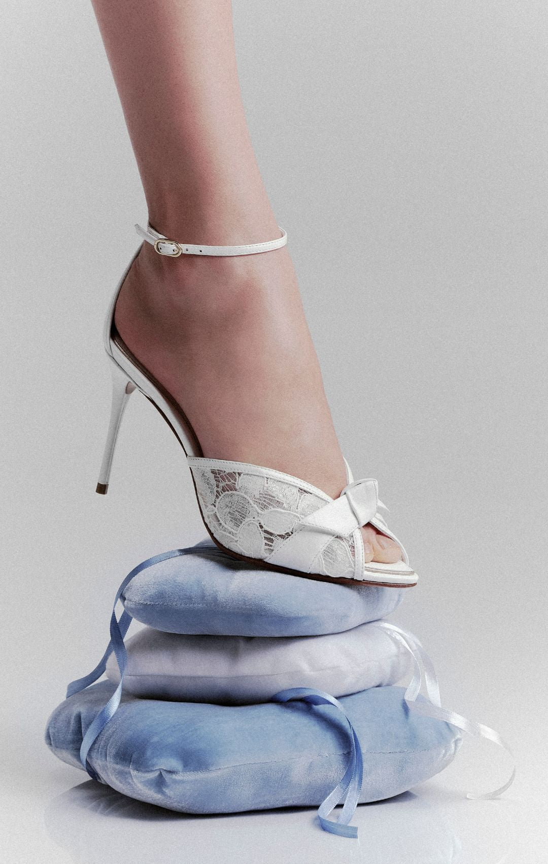 Close-up of the Clarita Bloom Lace 85 sandal balancing on stacked blue satin cushions, creating a delicate, whimsical fashion still life.