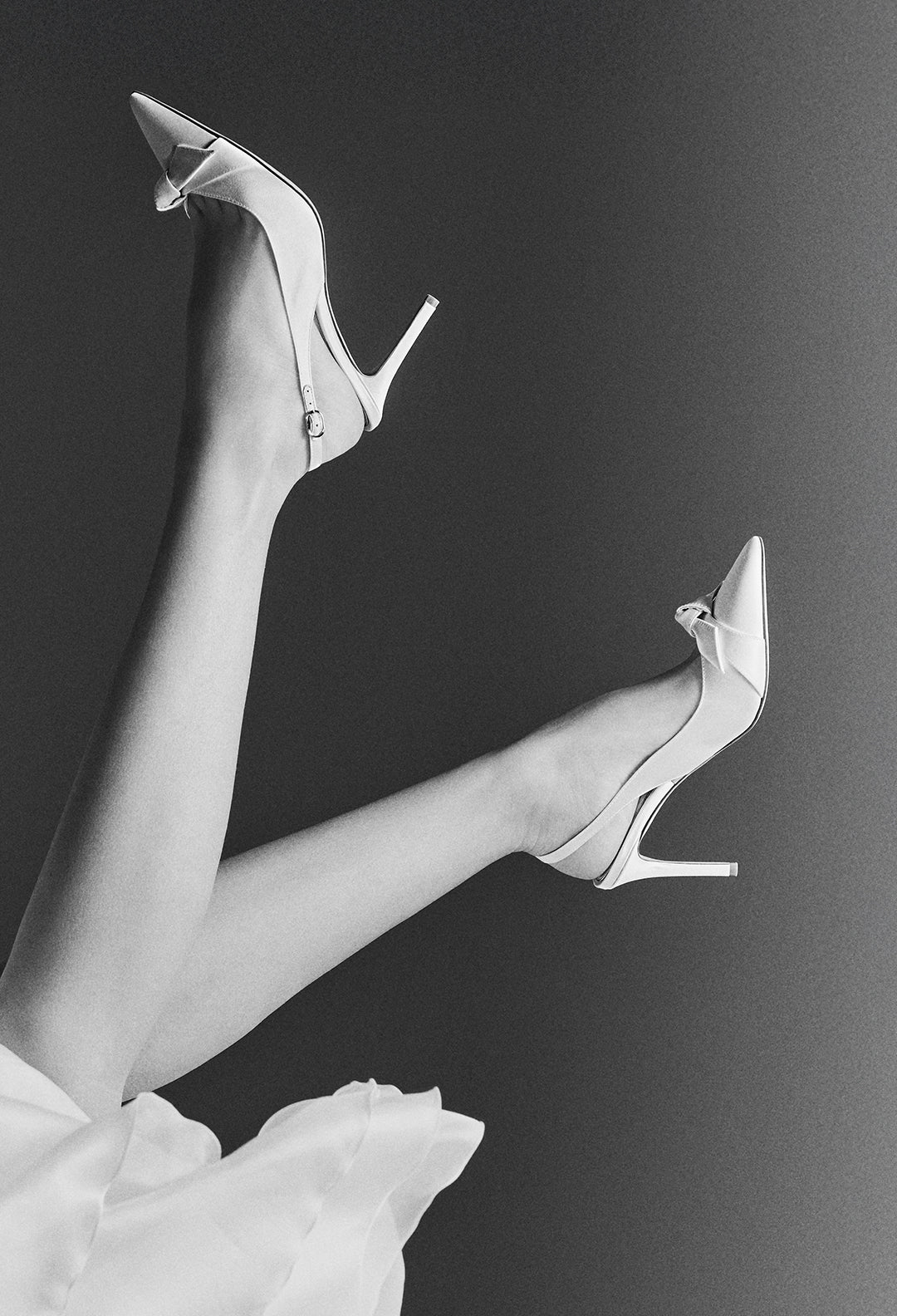 A black and white photo of a person's legs, crossed in the air, wearing elegant white pointed-toe heels with a bow detail.