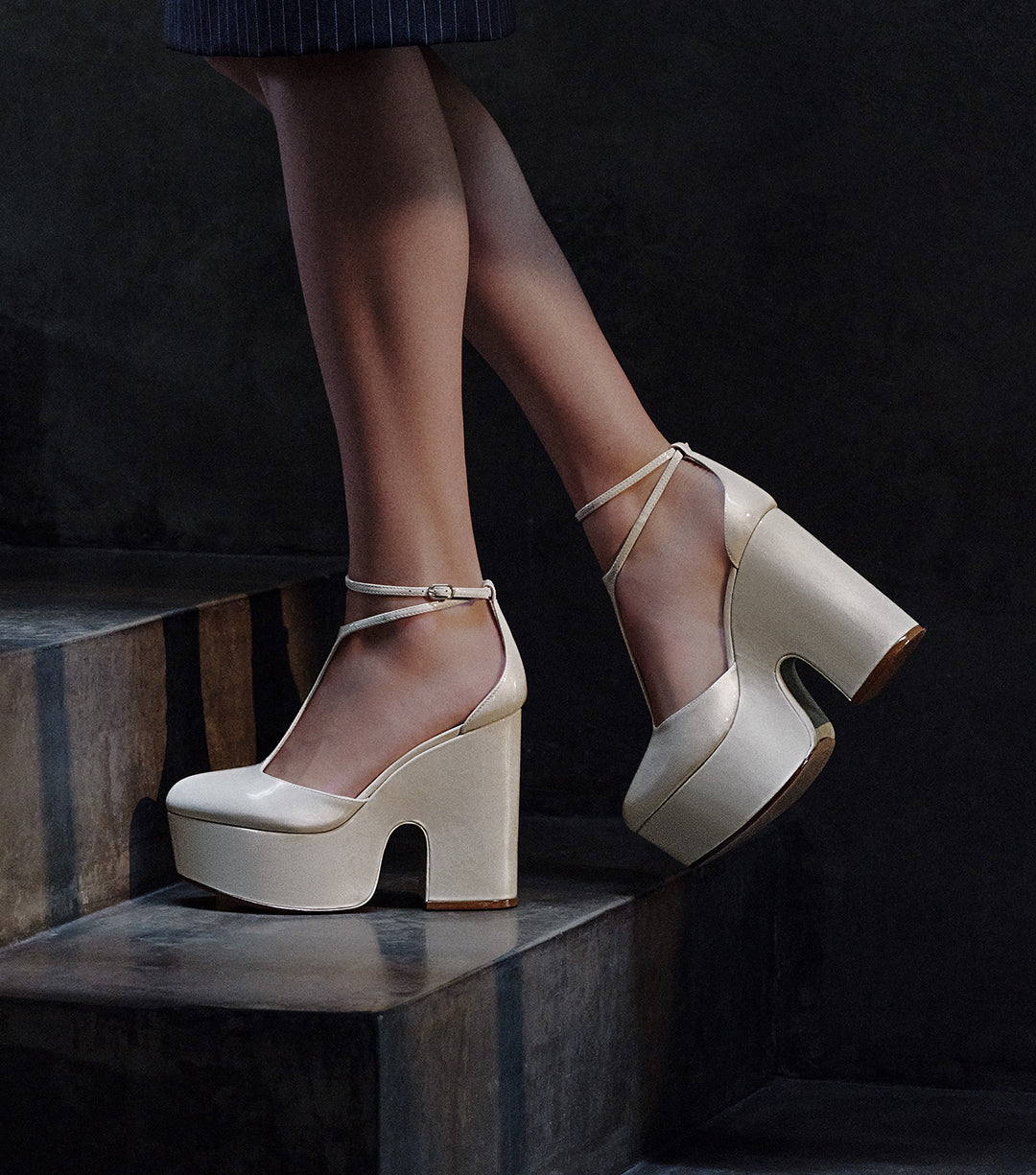 A close-up of a person's feet on a dark staircase, wearing a pair of high-platform ivory or white heels with an ankle strap.