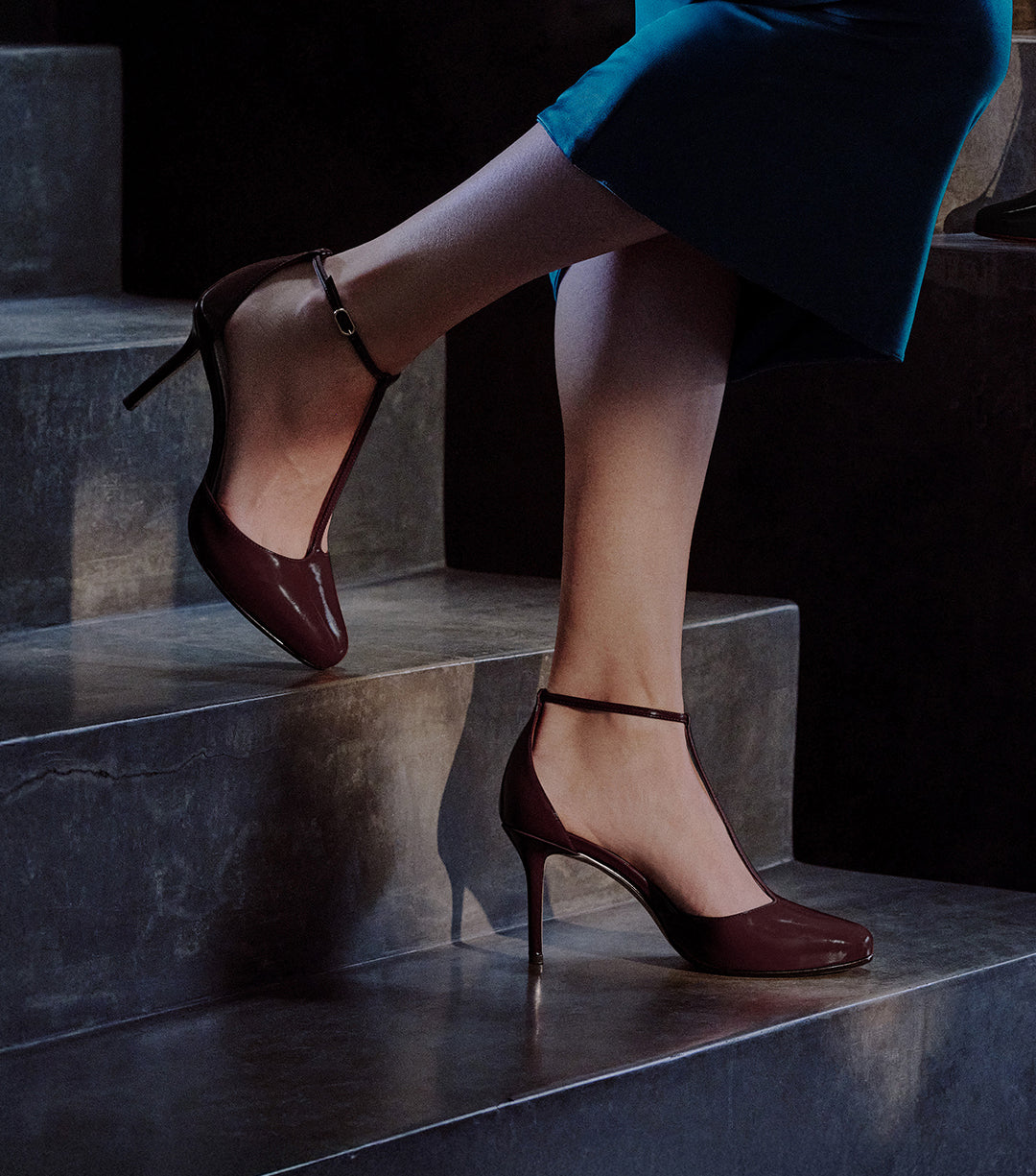 A person's legs walking up dark, concrete stairs, wearing glossy burgundy pumps with a pointed toe and a t-strap ankle strap.