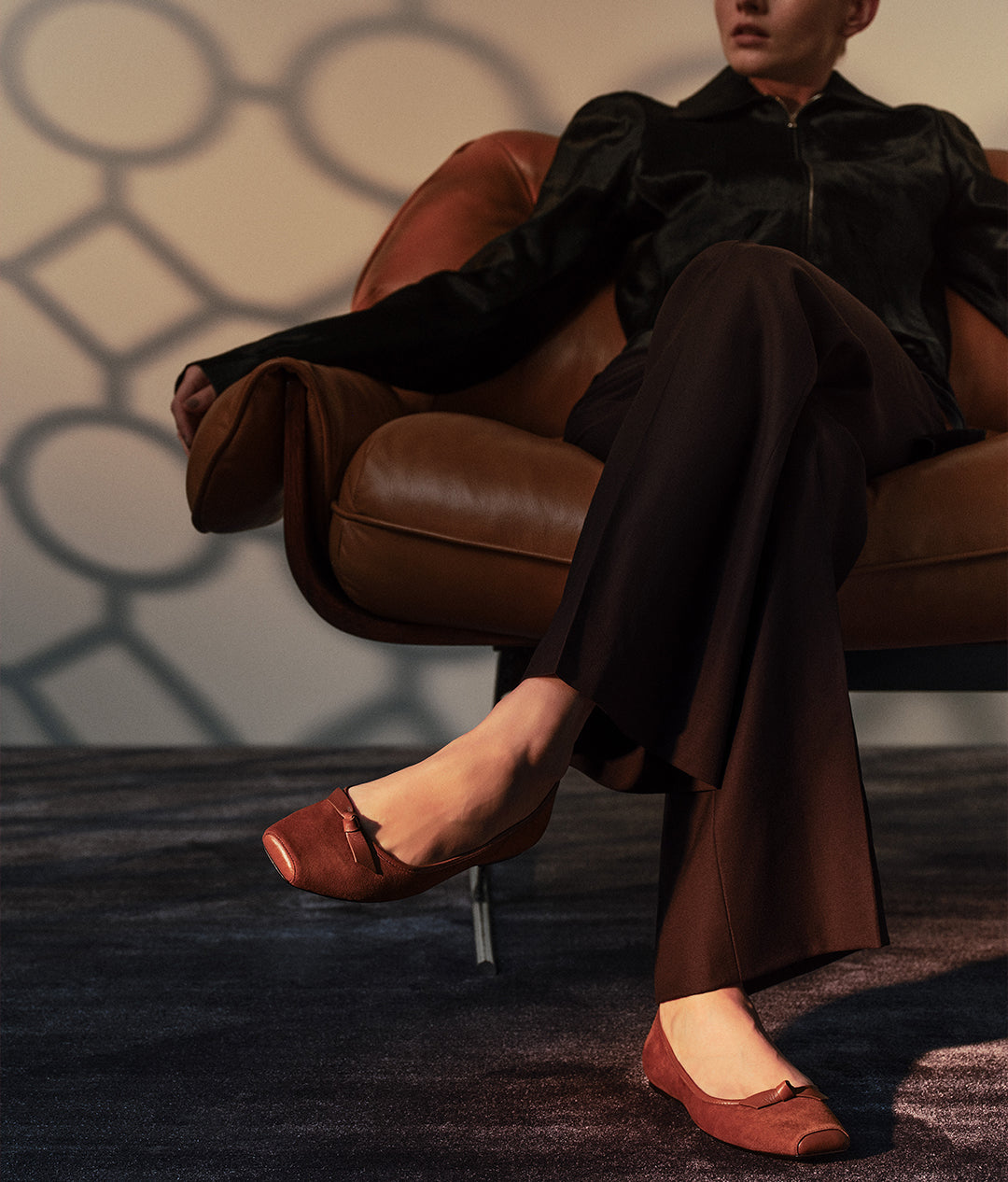  A person wearing a black jacket and brown pants, seated in a brown leather chair, with their feet in a pair of brown suede square-toe flats with a bow.
