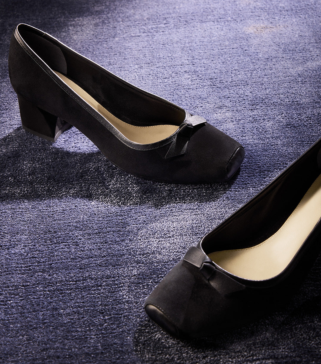 A pair of black suede pumps with a small block heel, a square toe, and a small bow detail at the front.