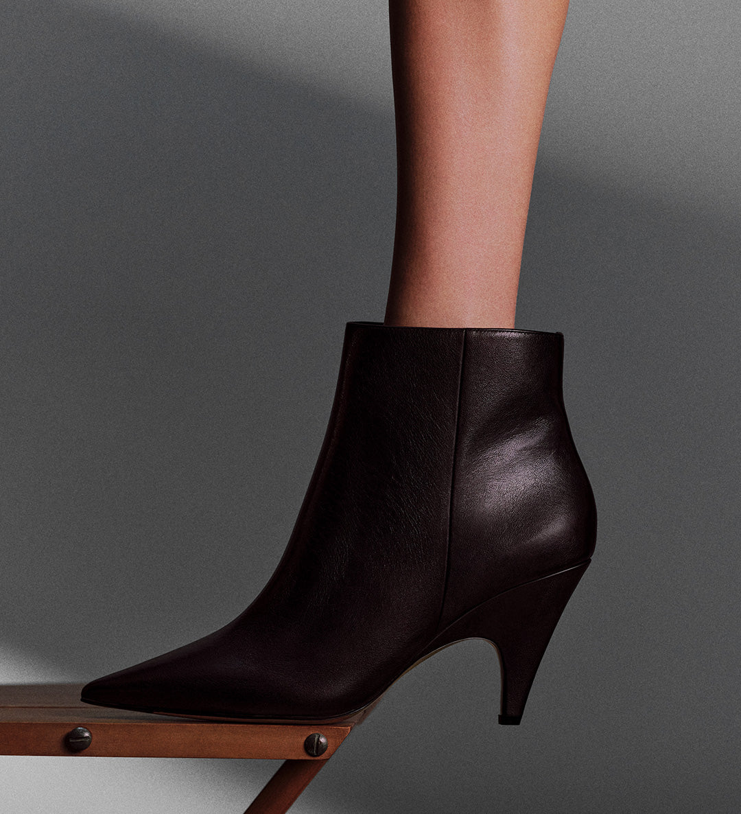 A close-up of a person's ankle and foot in a dark brown leather ankle boot with a pointed toe and a unique, geometric heel.