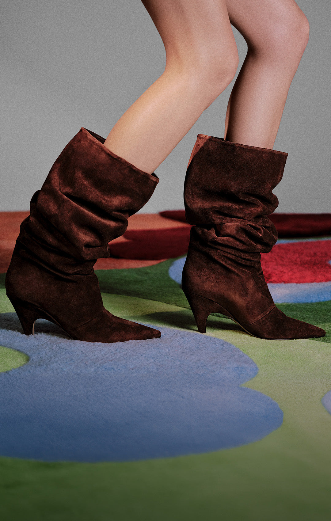  A person's legs are shown, wearing slouchy brown suede boots with pointed toes and kitten heels.