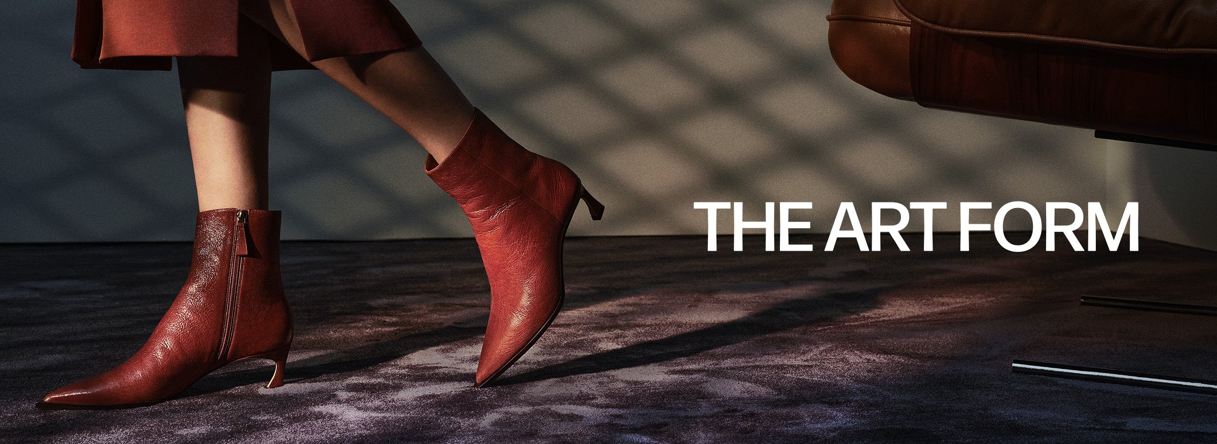  A close-up of two pairs of boots. One person is wearing black, textured ankle boots with a low heel and black leather pants. Another person is wearing similar red-brown ankle boots, with their feet crossed over the other person's lap. The text "THE ART FORM" is at the bottom of the image.