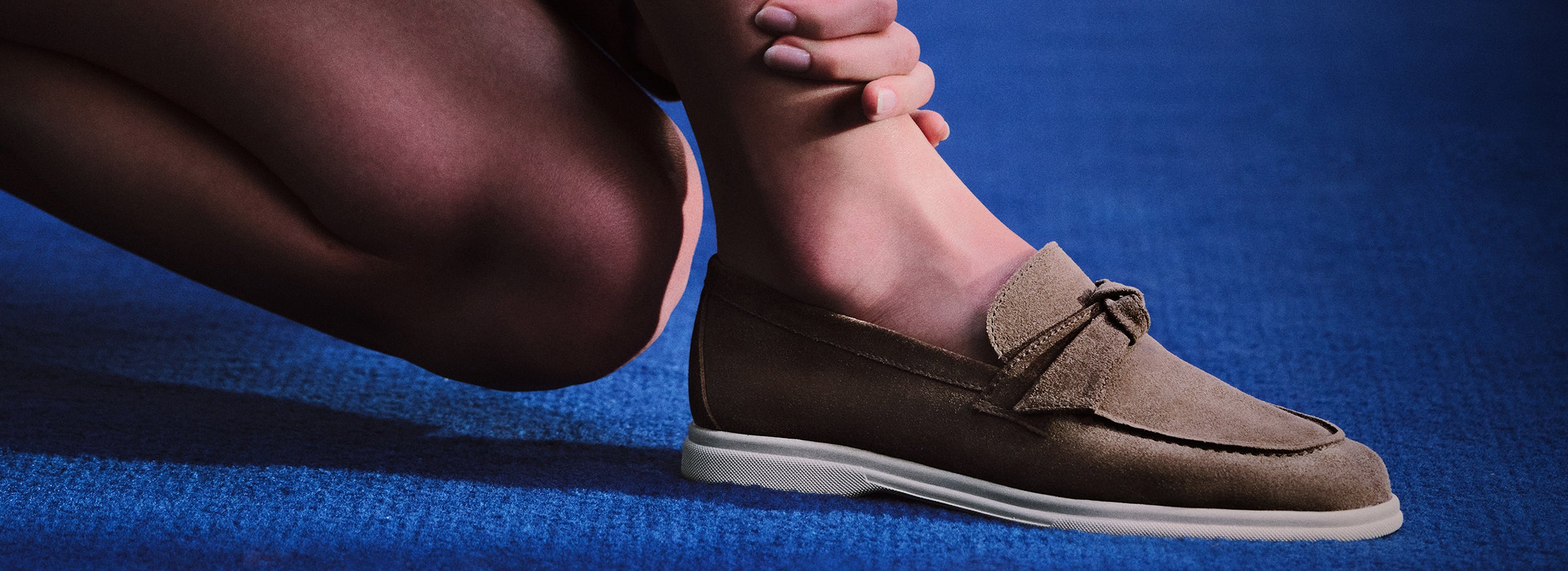  Close-up of a person's leg and foot wearing a brown suede loafer with a small bow, crouching on a bright blue carpet.