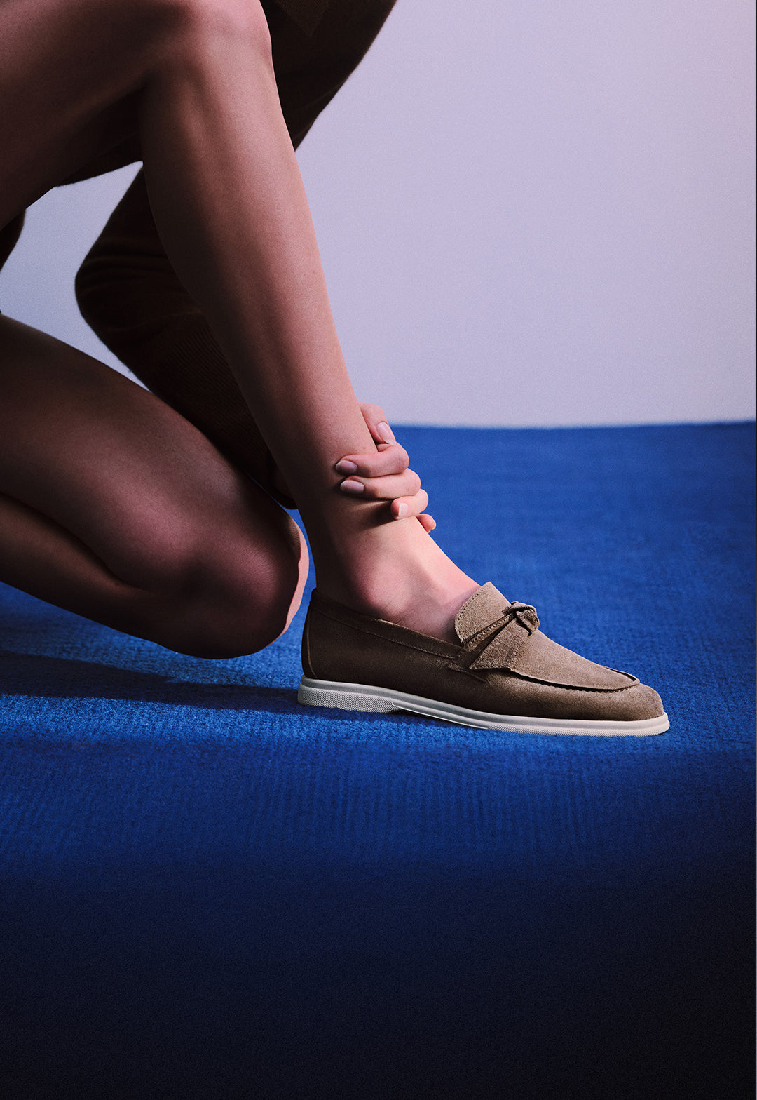  Close-up of a person's leg and foot wearing a brown suede loafer with a small bow, crouching on a bright blue carpet.