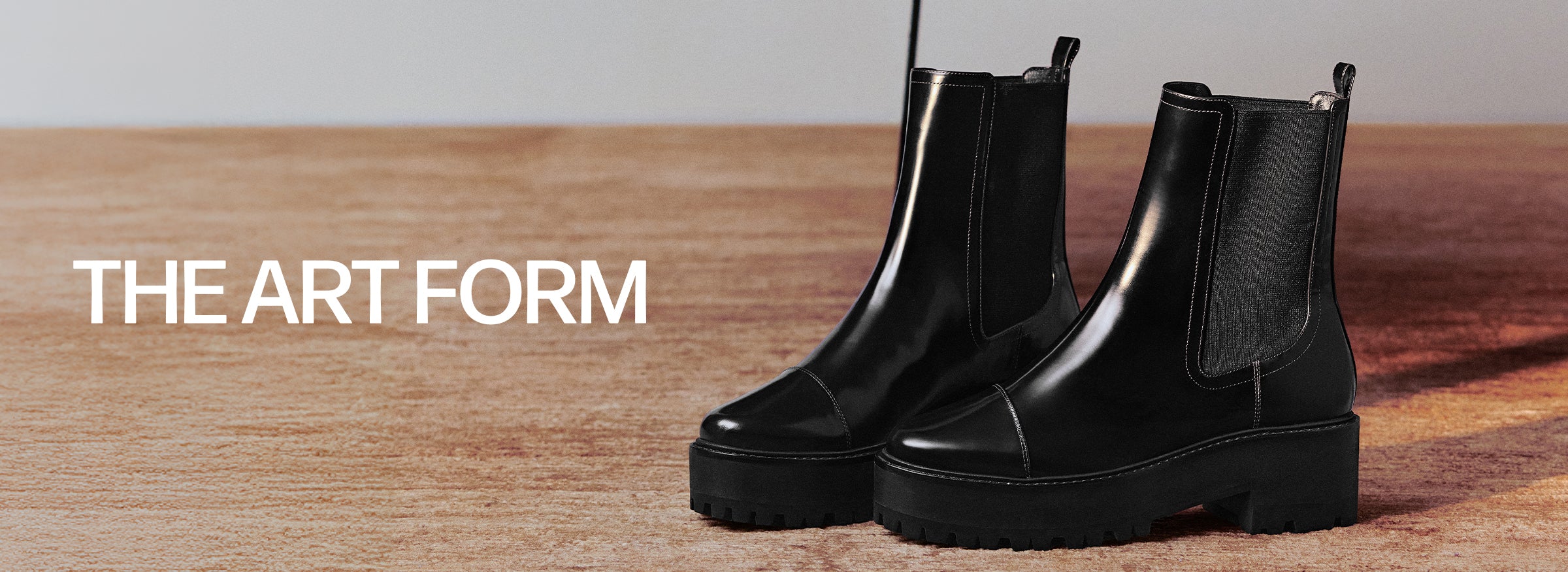  A pair of shiny, black leather Chelsea boots with chunky lug soles, placed on a light wooden floor. The text "THE ART FORM" is on the left.