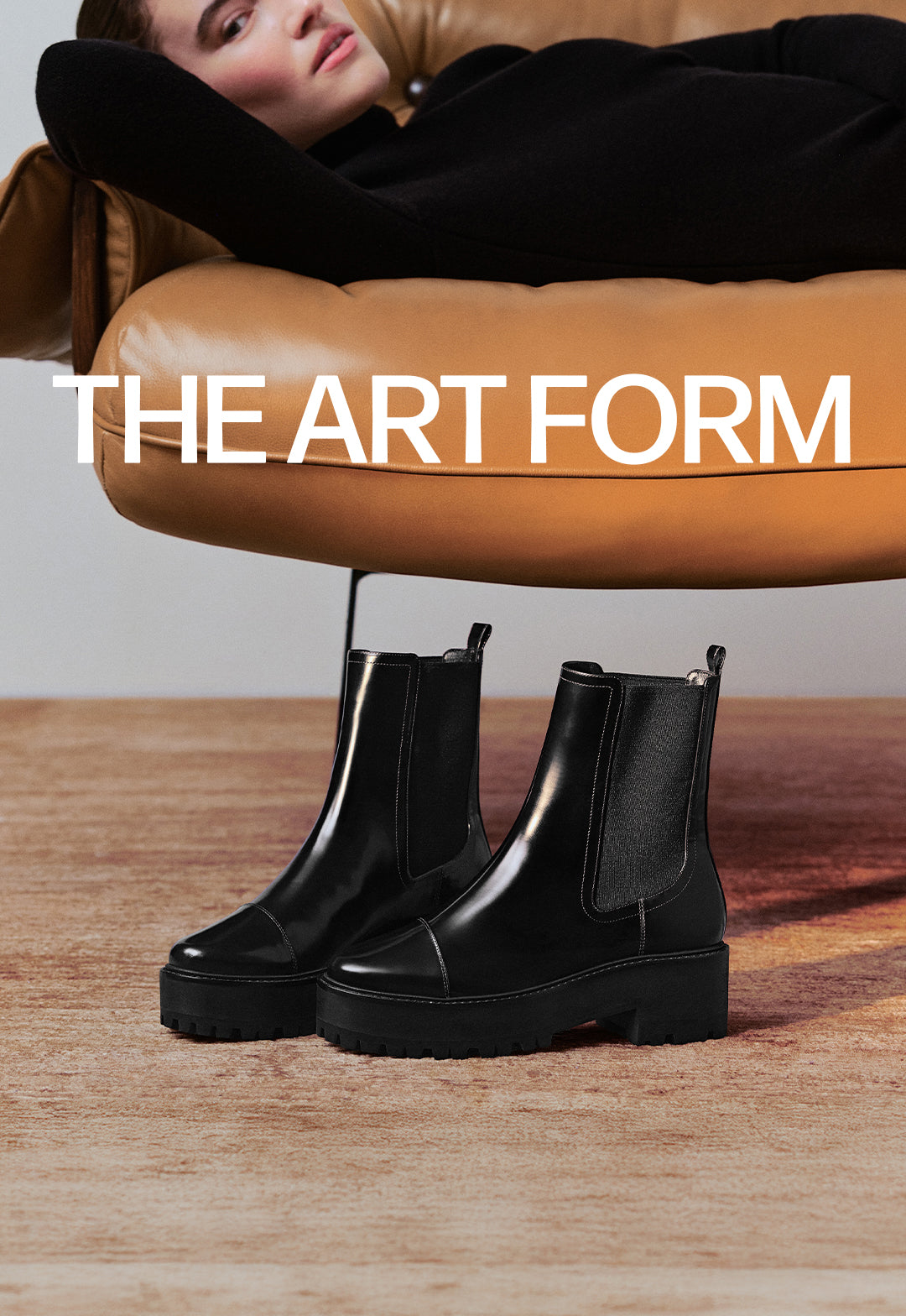  A pair of shiny, black leather Chelsea boots with chunky lug soles, placed on a light wooden floor. The text "THE ART FORM" is on the left.
