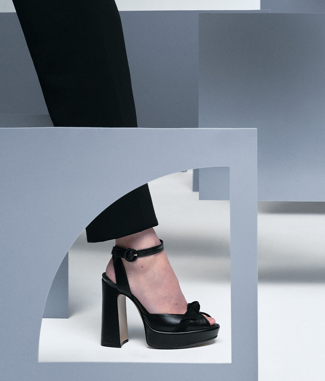  A close-up of a person's foot in a black leather platform sandal with a chunky heel and an ankle strap, stepping through a pale blue, geometric opening.