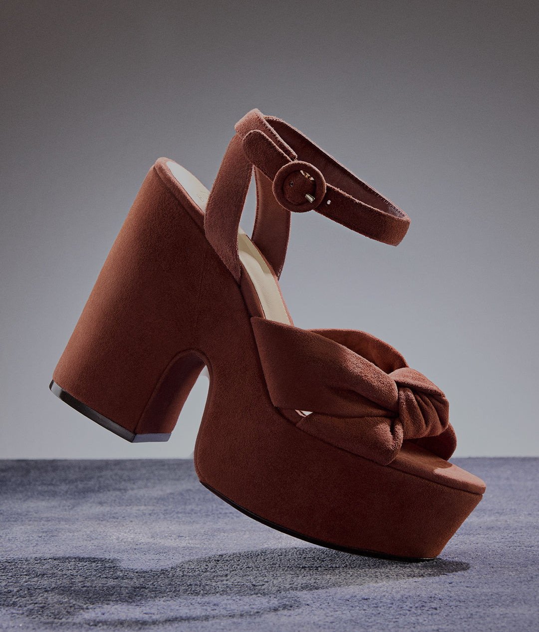  A chunky, terracotta-colored suede platform sandal with a thick block heel, an ankle strap, and a knotted bow detail across the toe. The shoe is suspended mid-air. Tap to Shop Sandals.