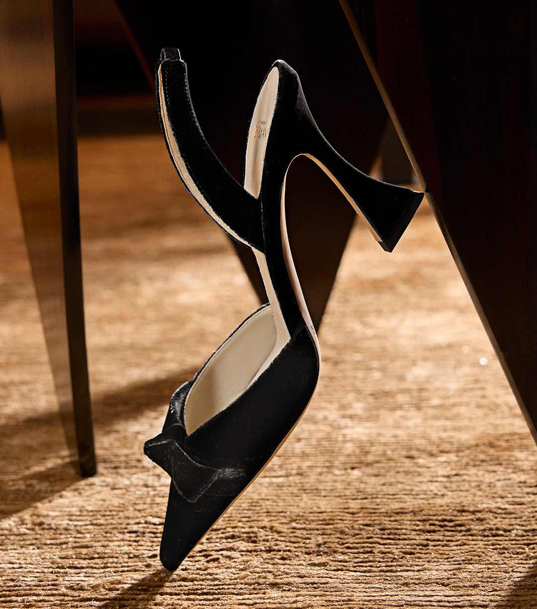 Pair of black velvet pointed-toe slingback pumps with sculptural heels and bow detail from Alexandre Birman’s New Pumps collection, displayed elegantly against a warm-toned carpet and wooden table.