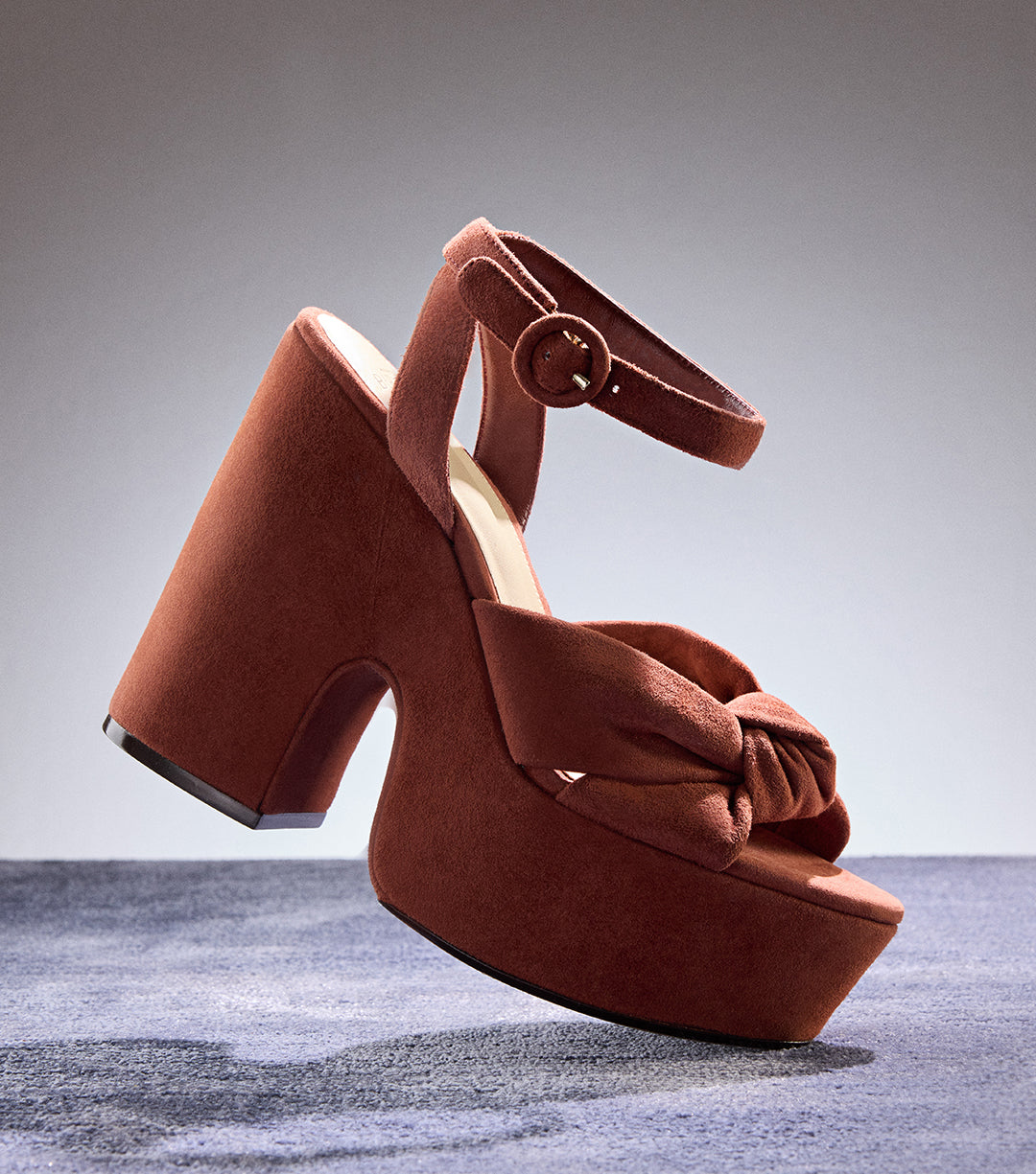 Pair of brickwood suede platform sandals with knotted front detail and ankle strap from Alexandre Birman’s New Sandals collection, displayed against a minimalist gray background highlighting their sculptural silhouette.