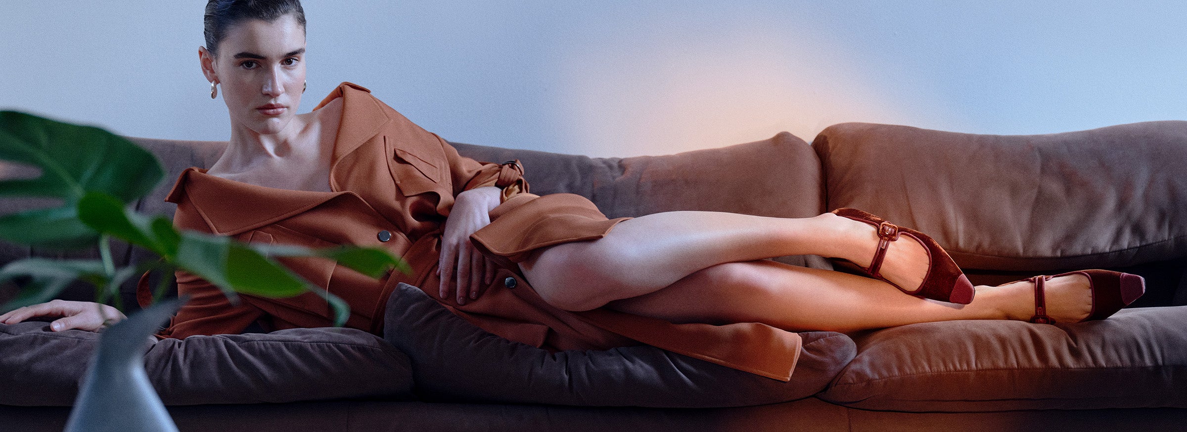  Model wearing brown trench dress and cuoio velvet ballet flats reclining on a sofa. The text "VELVET TOUCH" and the call to action "SHOP NOW" is to the bottom left of the image.