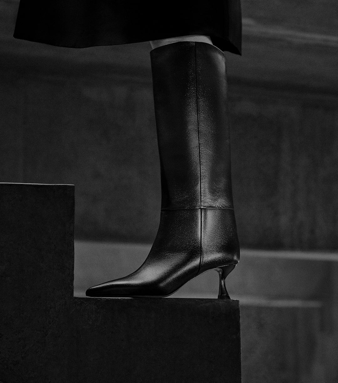 Black leather knee-high boots with pointed toes and sculptural geometric heels, shown on a model stepping up a staircase. Text on image reads ‘New Boots'.