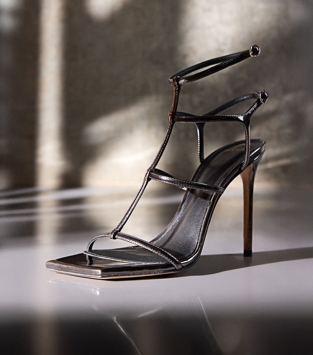 Elegant high-heeled sandal in specchio grafite leather with thin straps, displayed under soft natural light. Text on image reads ‘New Sandals.