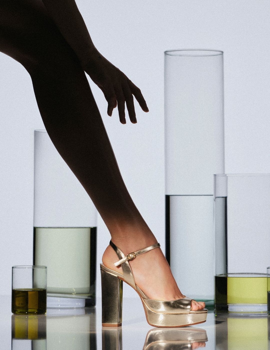A woman in sheer black tights reaches forward in a gold high-heeled sandal, standing among glass cylinders of water; the minimalist scene feels poised and elegant. Tap to shop