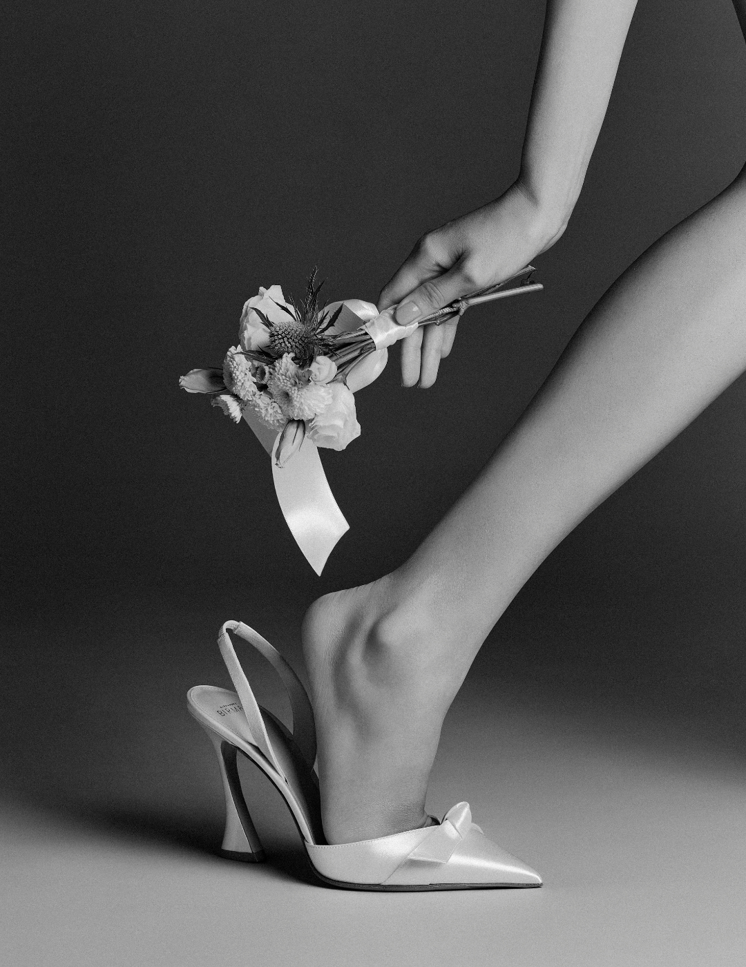 A monochrome image shows a person adjusting a high-heeled shoe on their foot while holding a delicate bouquet with ribbon, evoking elegance and grace.