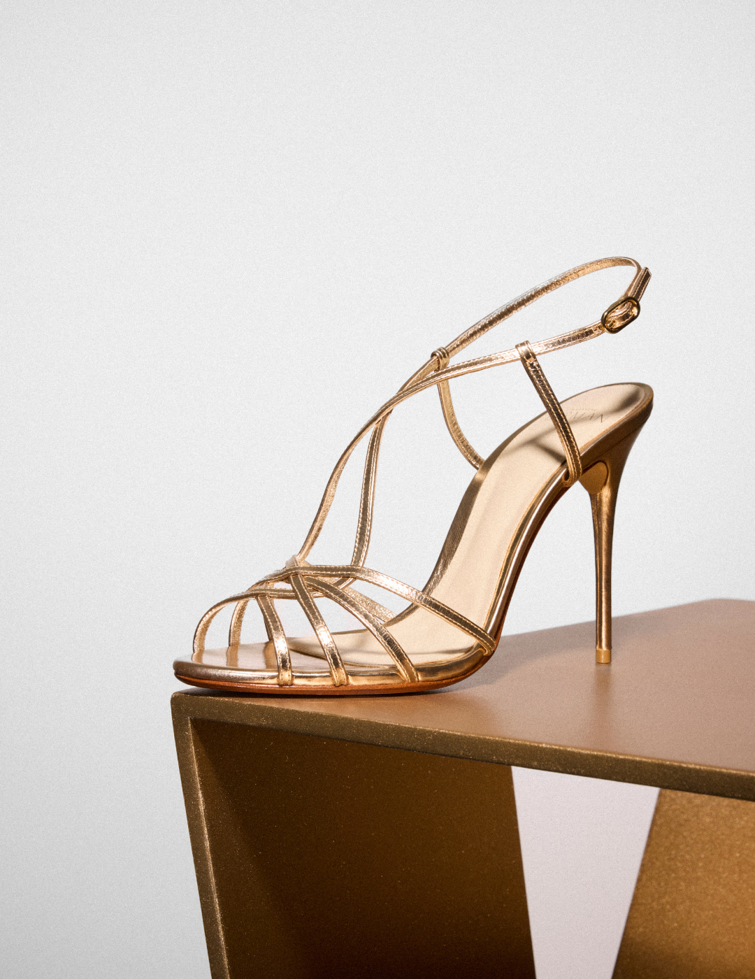 Elegant gold stiletto sandal with slender straps and open toe. Positioned on a golden geometric stand against a light background. Tap to shop