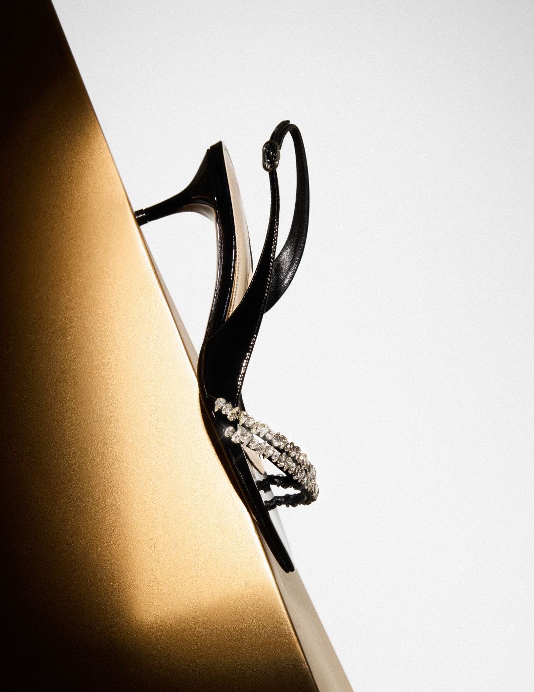 Black crystal-embellished slingback sandal displayed against a sculptural gold surface and white background. Tap to shop