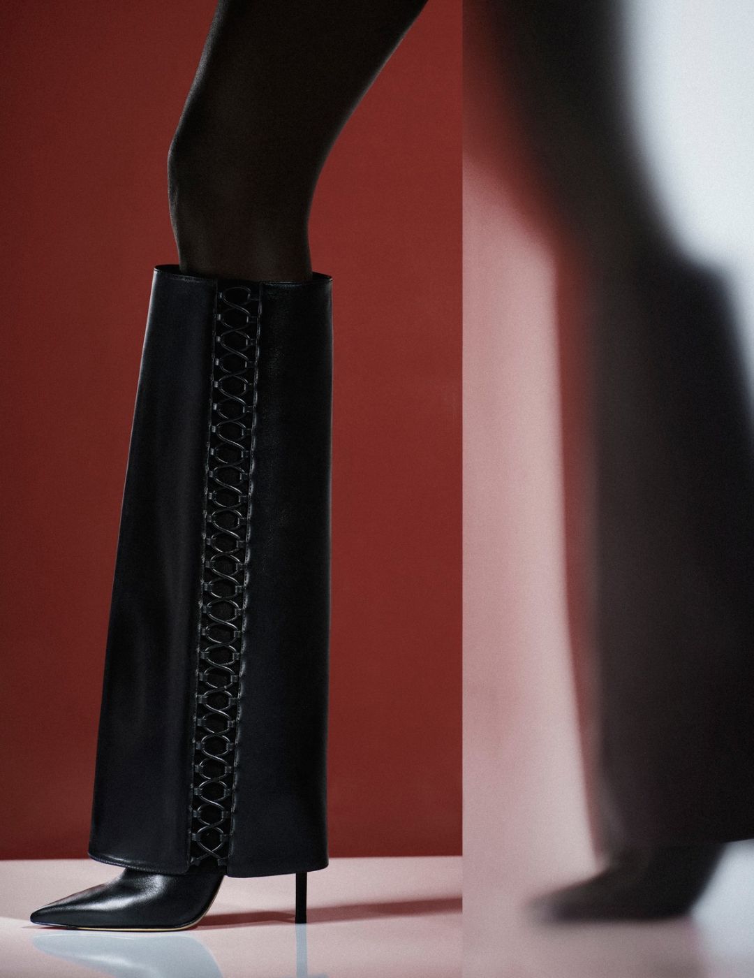 Black leather knee-high boots with decorative front lacing, modeled against a deep red backdrop; a blurred reflection adds a dramatic, fashion-editorial mood. Tap to shop.