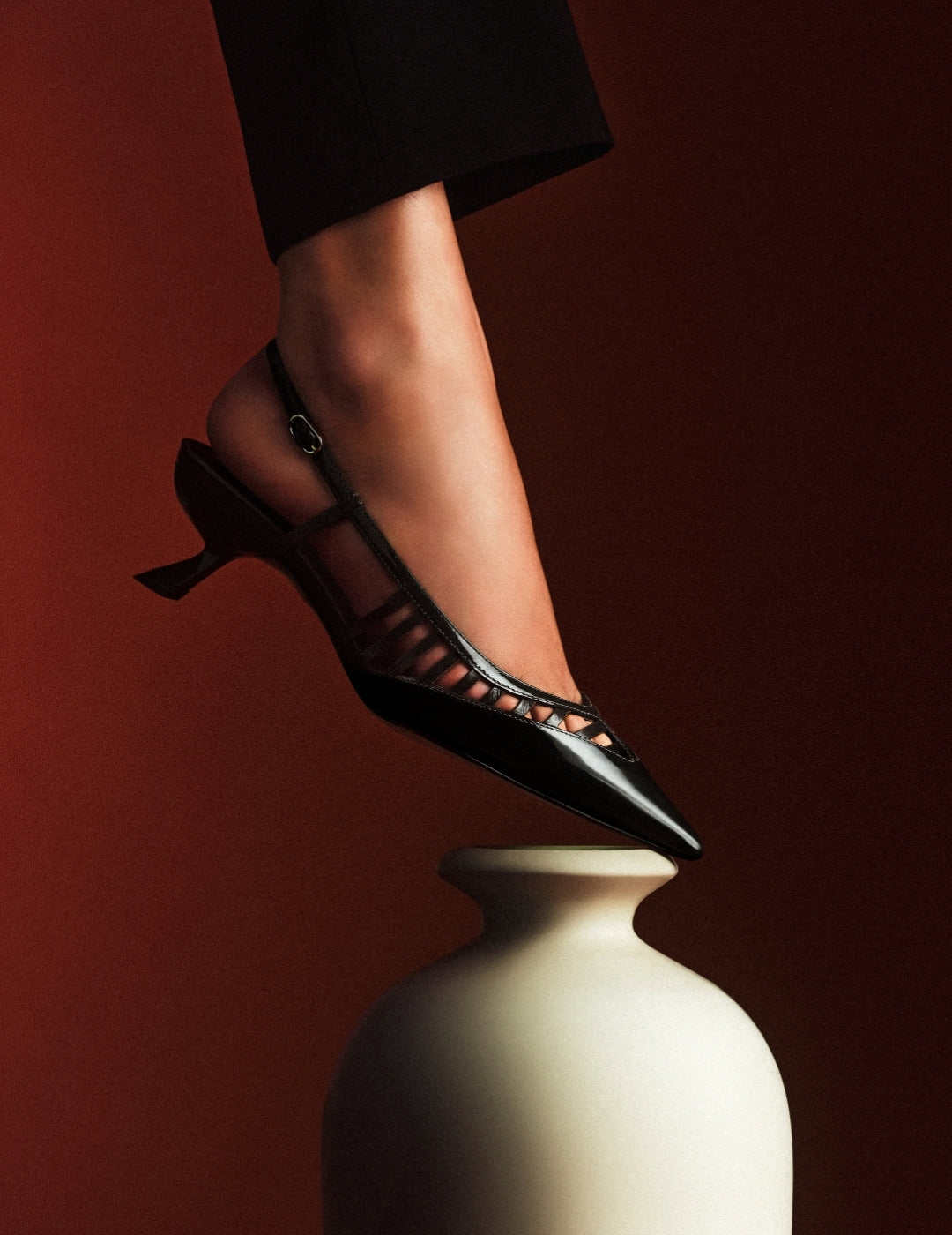 A foot in a black strappy high-heeled sandal balances on a cream ceramic vase against a deep red background, creating a dramatic fashion still life. Tap to shop.