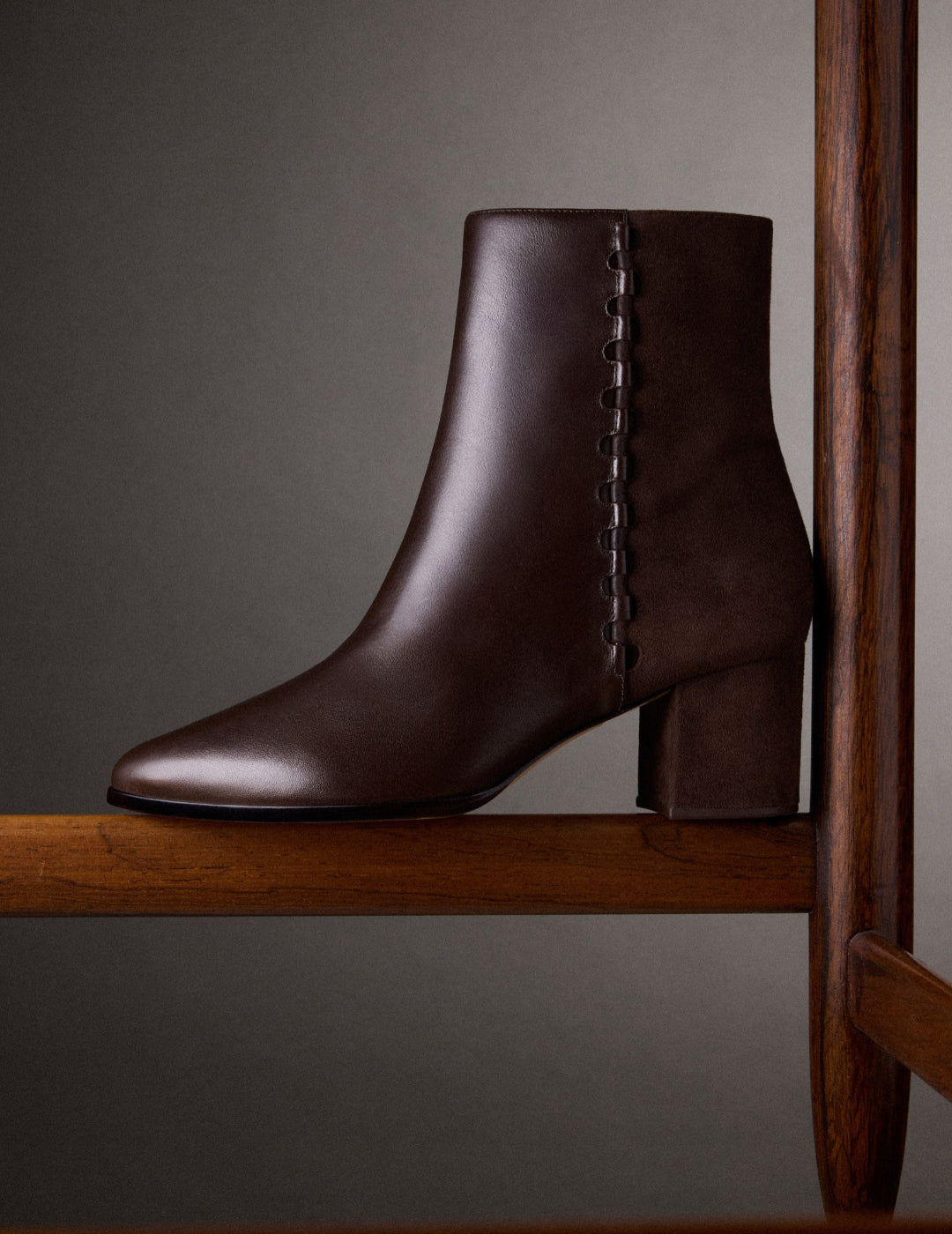 A polished dark-brown leather ankle boot with a chunky block heel and decorative side buttons rests on a wooden chair, evoking quiet sophistication. Tap to shop.