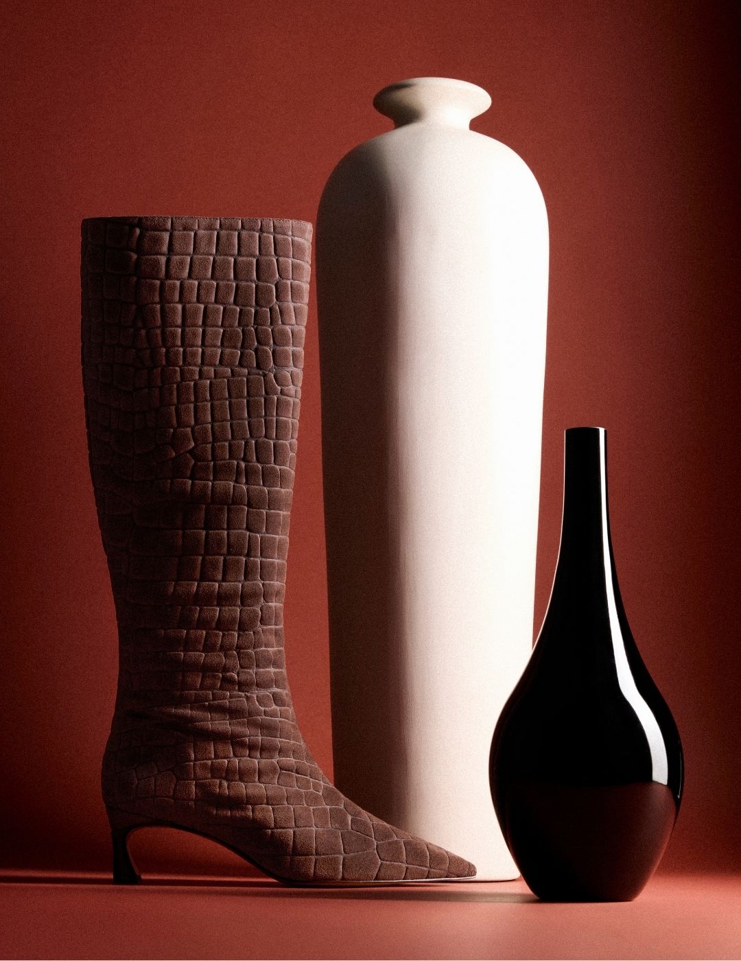 A textured brown knee-high boot, tall white ceramic vase, and glossy black bottle stand against a deep red backdrop, creating a dramatic still life. Tap to shop