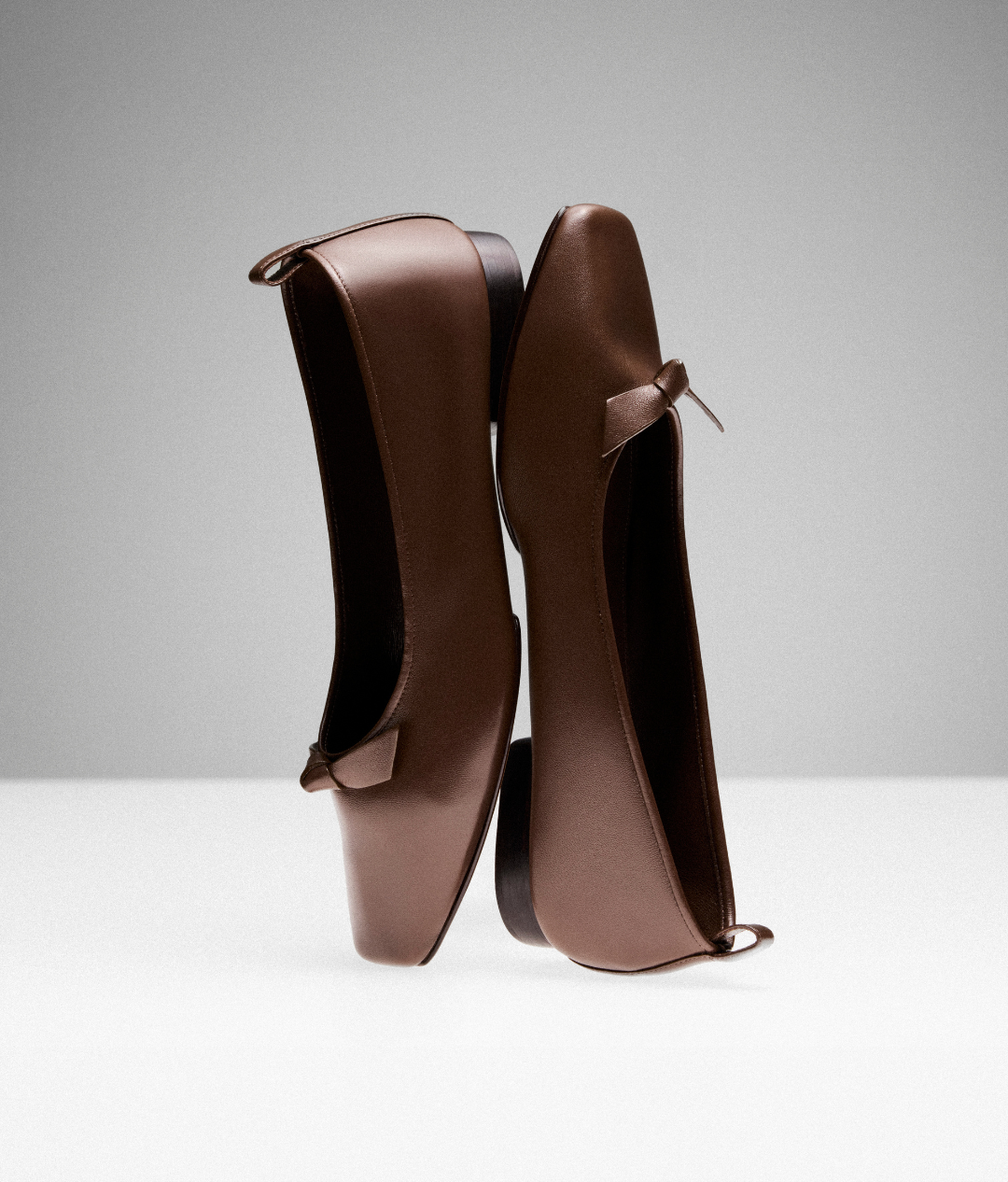 Brown leather ballet flats with small bows, creatively balanced heel to toe on a light surface. The setup conveys elegance and simplicity. Tap to shop