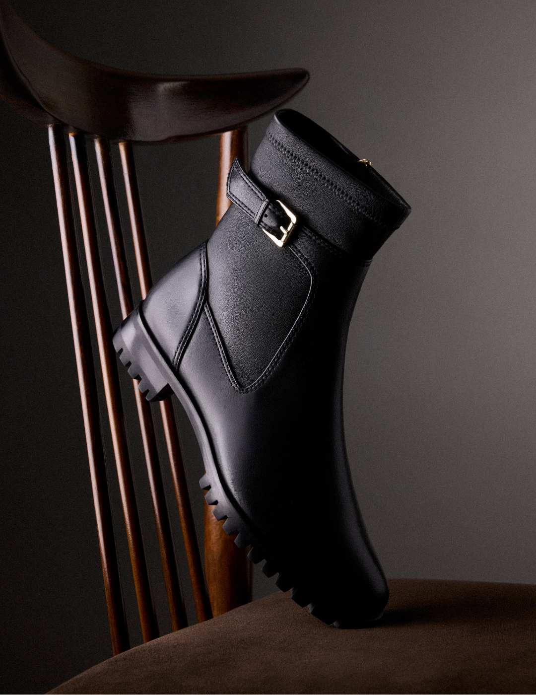 Black leather ankle boot with side buckle and rugged lug sole rests against a dark wooden chair, creating a sleek, dramatic studio mood. Tap to shop