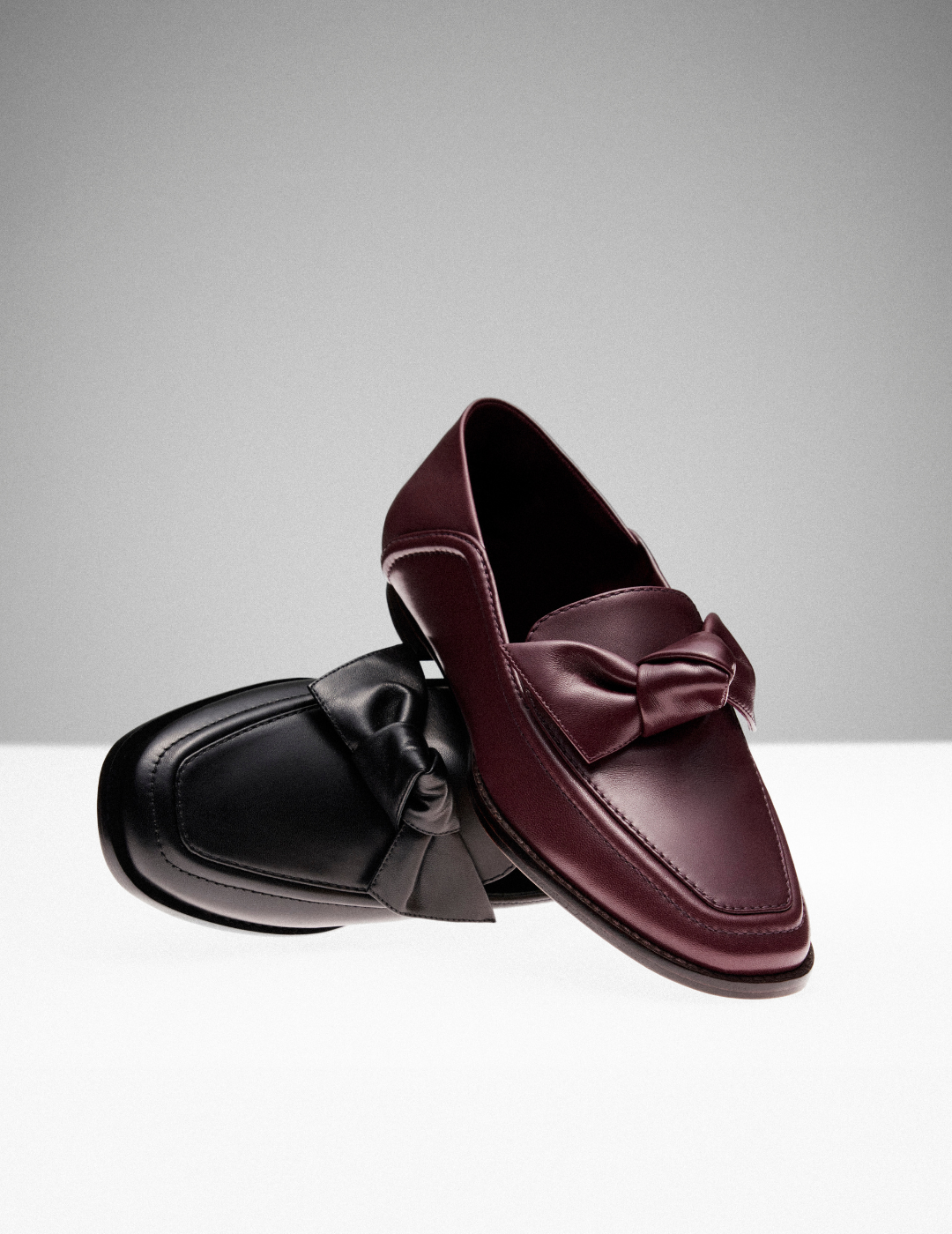 Two elegant leather loafers are displayed, one in deep burgundy and the other in black, each adorned with a stylish bow, against a minimalist backdrop. Tap to shop