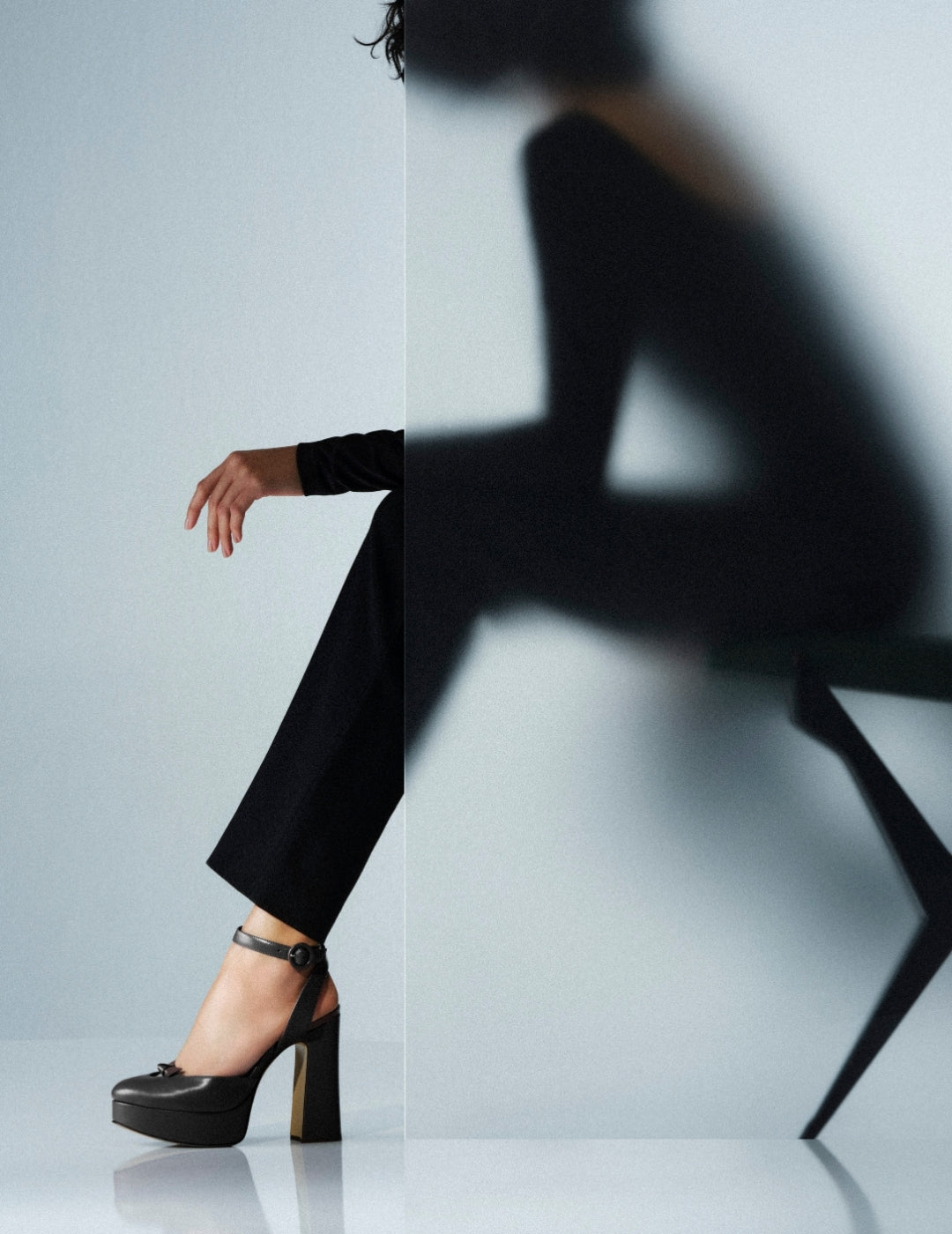  Fashion editorial image of a woman in black trousers and platform heels, split with a blurred seated silhouette against a minimalist backdrop. Tap to shop
