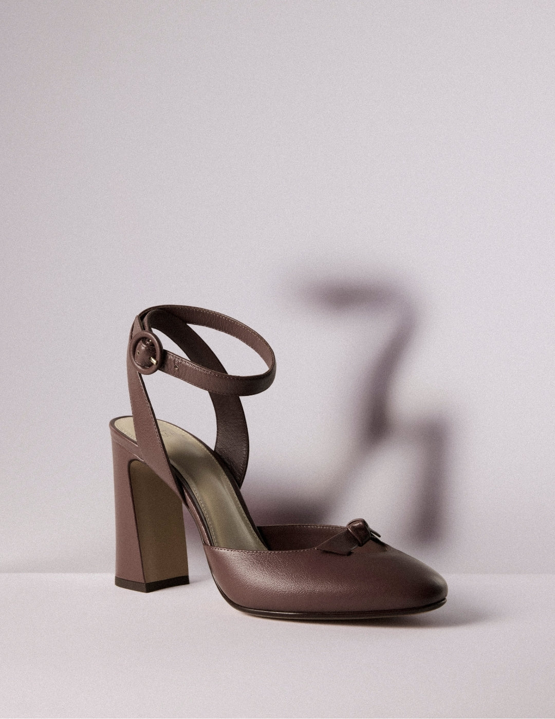 Brown leather Mary Jane high-heeled shoe with ankle strap on a pale surface, casting a soft shadow in an elegant, minimalist studio scene. Tap to shop.