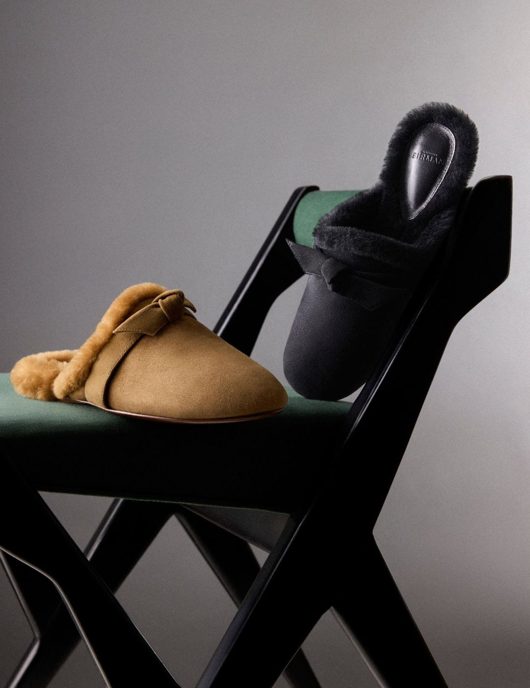 Two cozy suede slippers—one tan and one black with plush lining—rest on a black folding chair against a gray backdrop, creating a calm, inviting mood.ap to shop