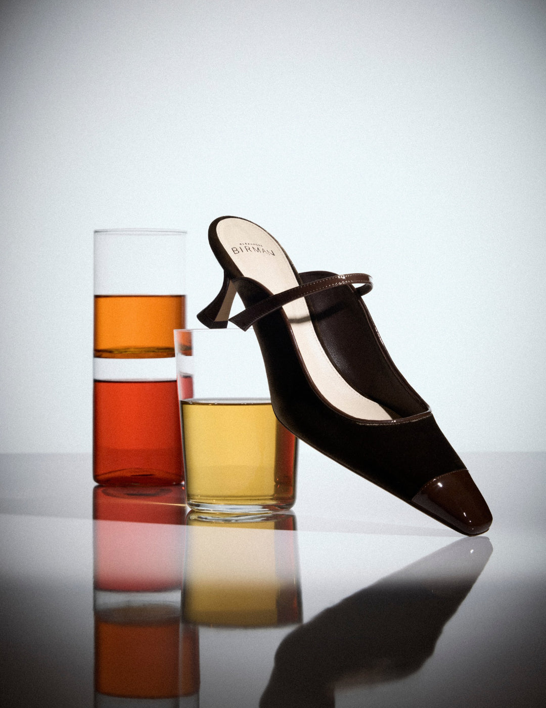  A dark brown, open-toe slingback heel leans across three glasses of amber, orange, and golden drinks, reflected on a glossy surface; sleek, upscale mood. Tap to shop