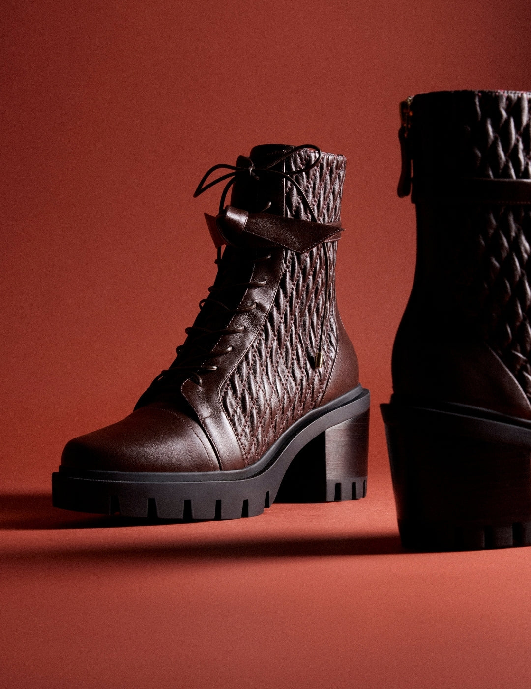  Two dark brown quilted leather ankle boots with chunky lug soles stand against a warm red backdrop, creating a bold, dramatic fashion mood. Tap to shop
