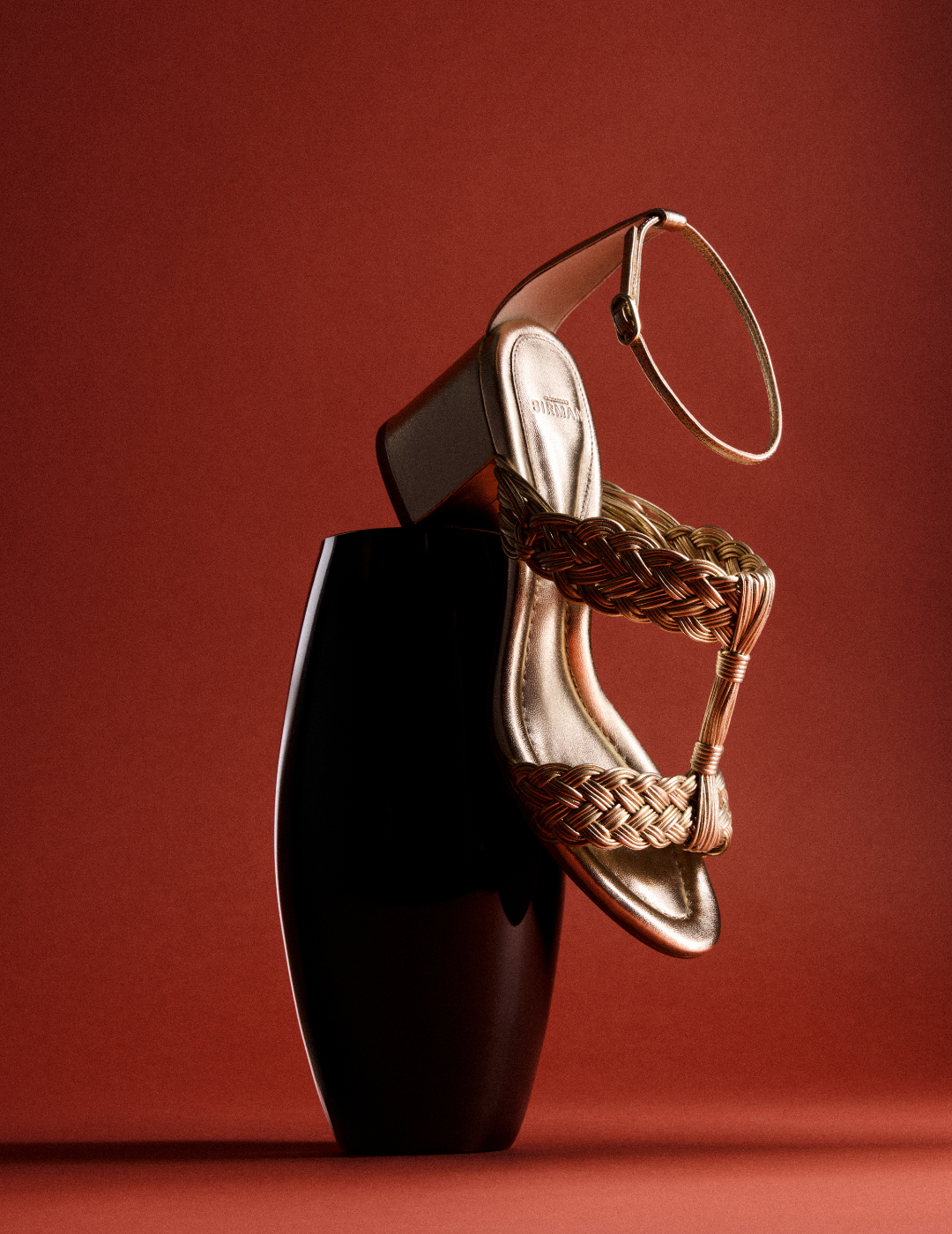 A sculptural gold strappy high-heeled sandal rests on a glossy black pedestal against a deep red background, creating a luxurious mood. Tap to shop