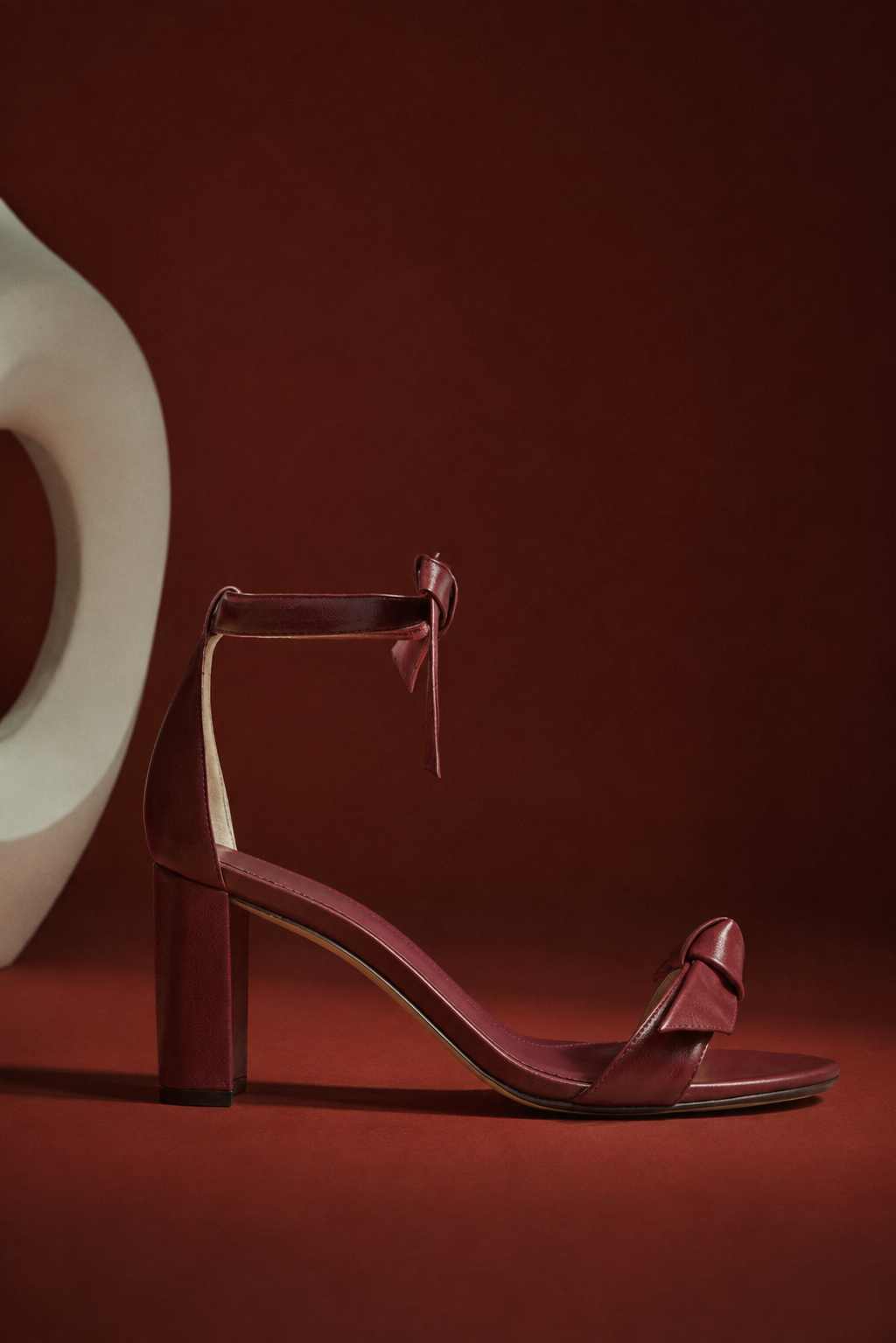 Elegant burgundy leather wedge sandal with ankle strap, sculpted heel, and toe bow, displayed against a rich red backdrop in dramatic light. Tap to shop