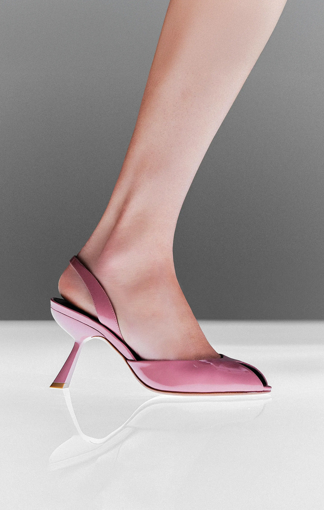  Model wearing a pink patent leather peep-toe slingback pump with a sculptural heel against a gray background.