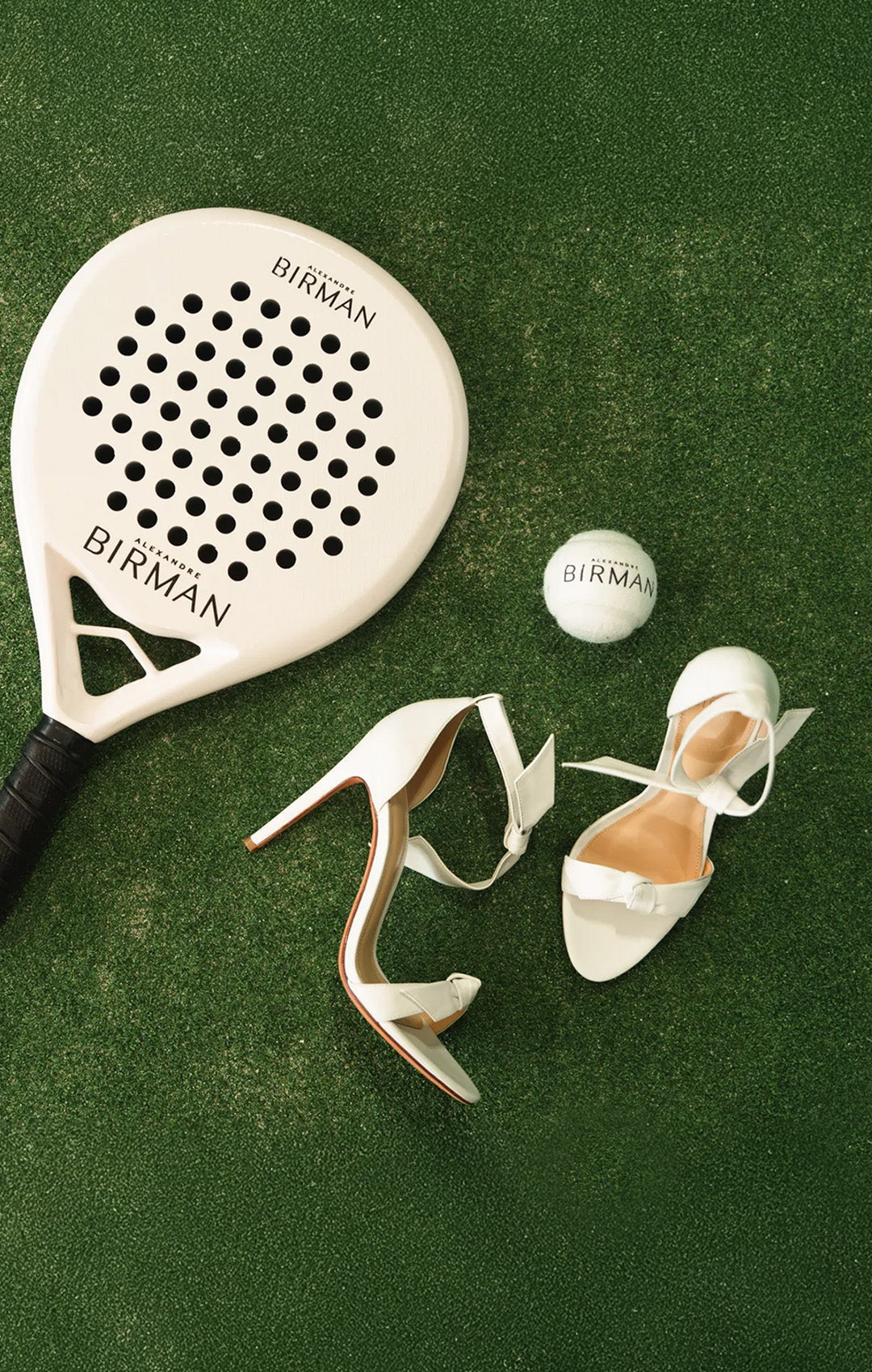  White Alexandre Birman Clarita sandals beside a branded padel racket and ball on a green court.