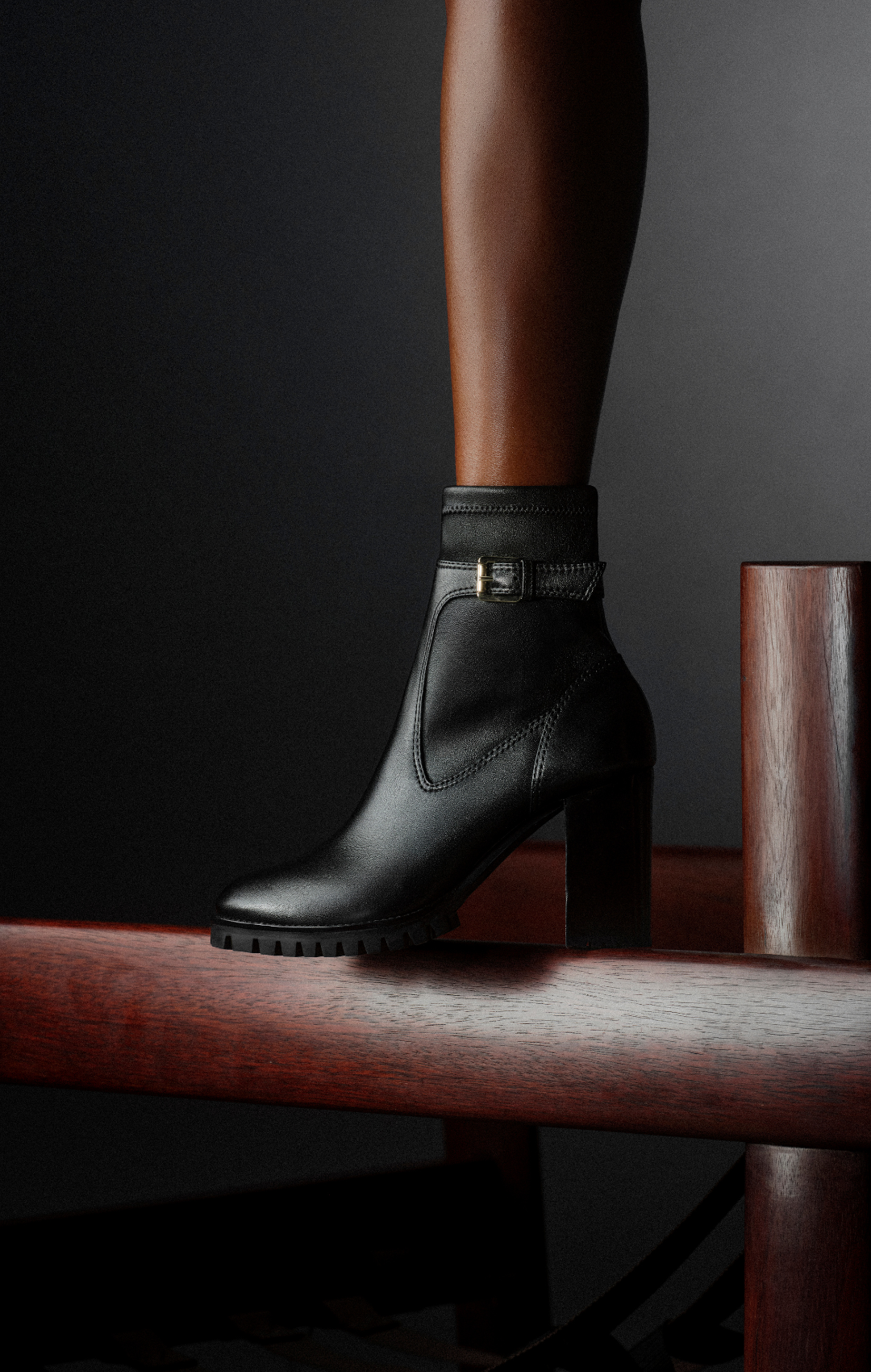 Black leather ankle boot with a high block heel rests on a wooden rail against a dark background. Shop Now.